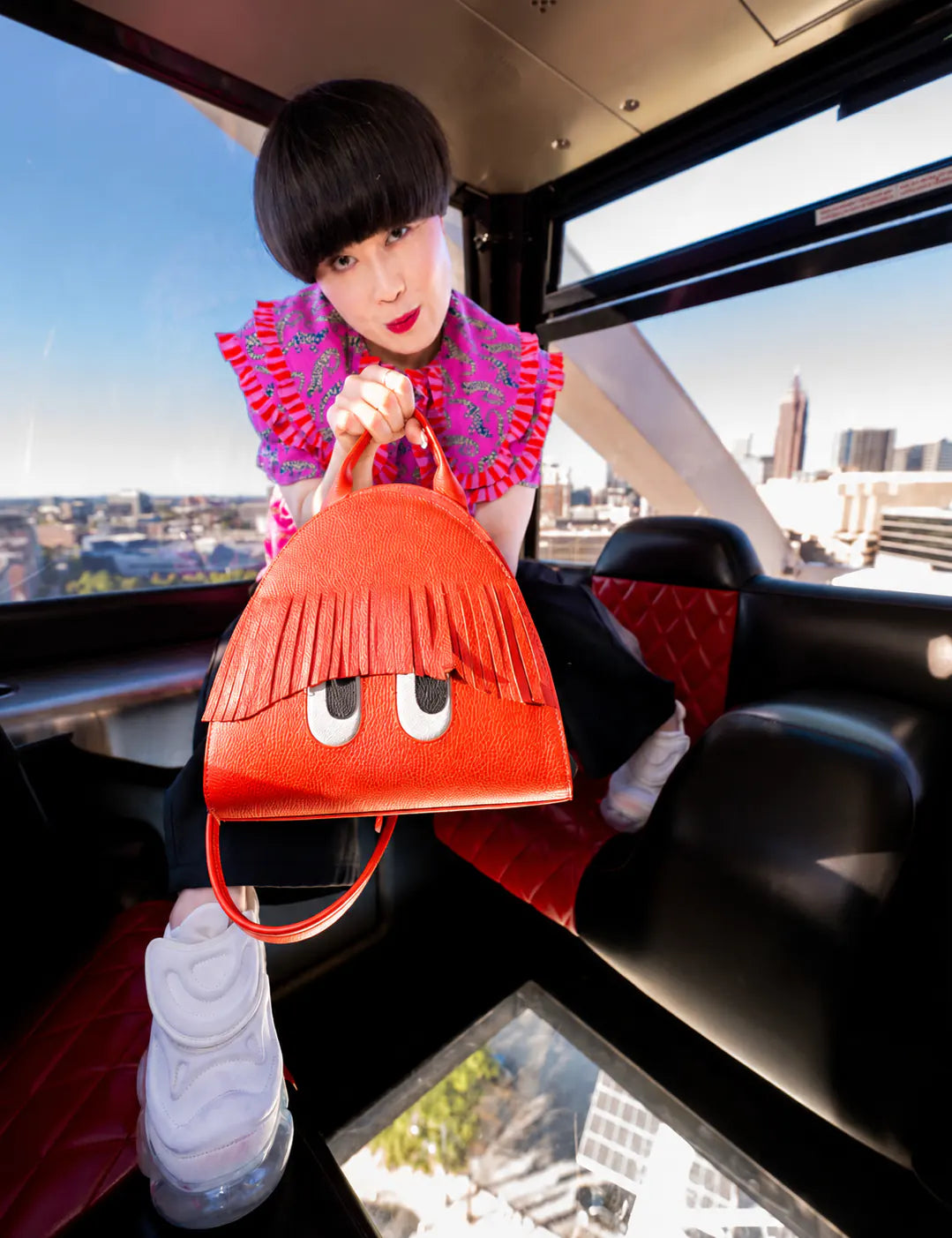 Model in a vibrant pink patterned top showcases an orange Min & Mon handbag with playful fringe bangs and engraved eyes while posing inside a ferris wheel car overlooking the city.