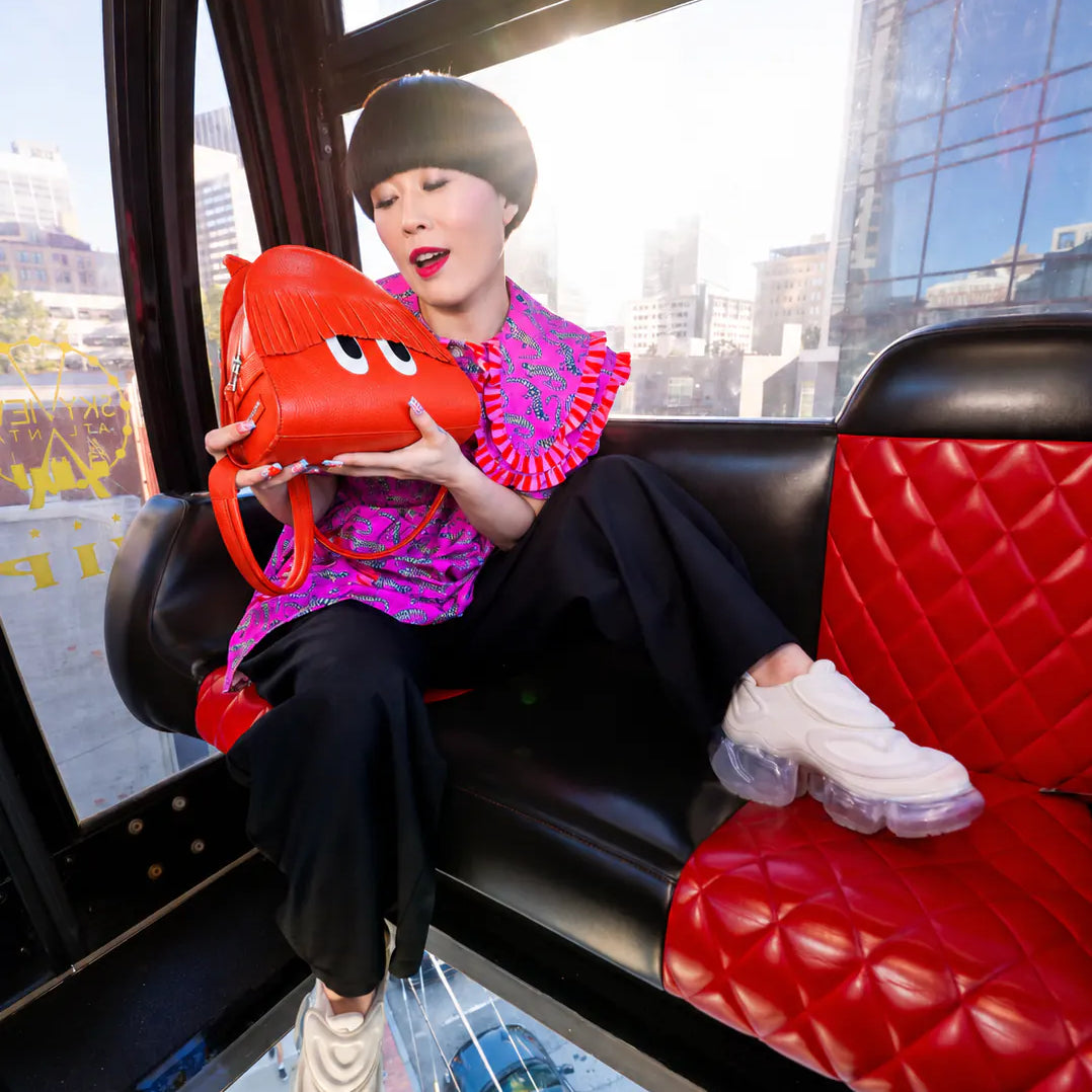 A woman sits on a red quilted leather seat holding a bright red cartoon-style shoulder bag with white eyes. She wears a pink and purple patterned top, black pants, and white sneakers, with a city street visible through the bus windows.