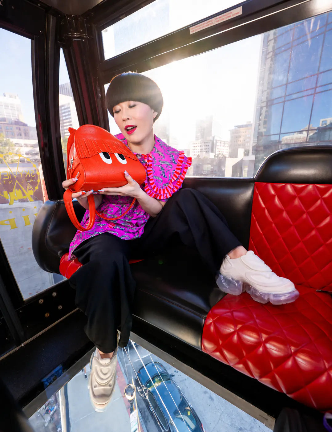 A woman sits on a red quilted leather seat holding a bright red cartoon-style shoulder bag with white eyes. She wears a pink and purple patterned top, black pants, and white sneakers, with a city street visible through the bus windows.