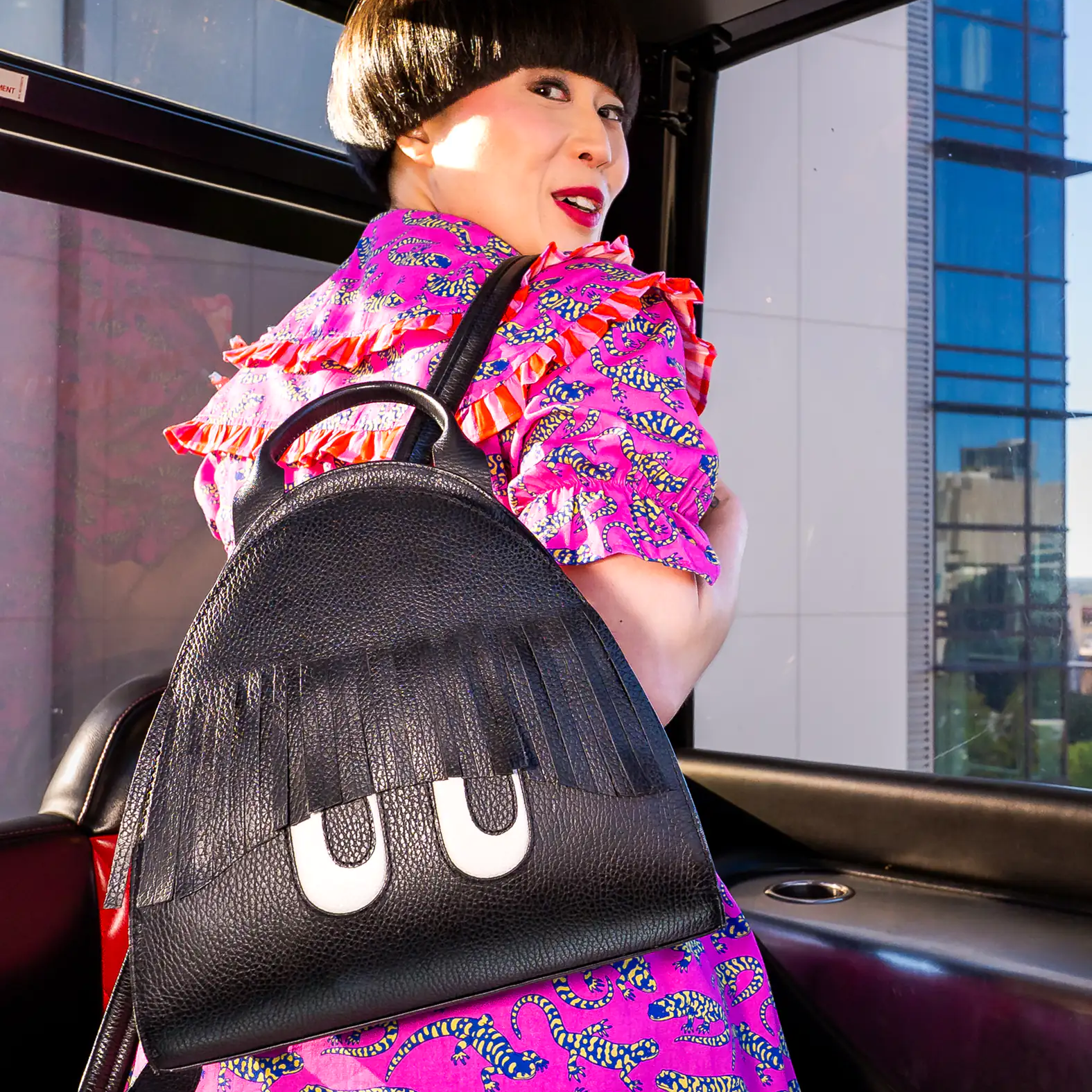Model in a vibrant pink patterned outfit poses inside a ferris wheel, showcasing a black Min & Mon backpack featuring playful fringe bangs and engraved cartoon eyes.