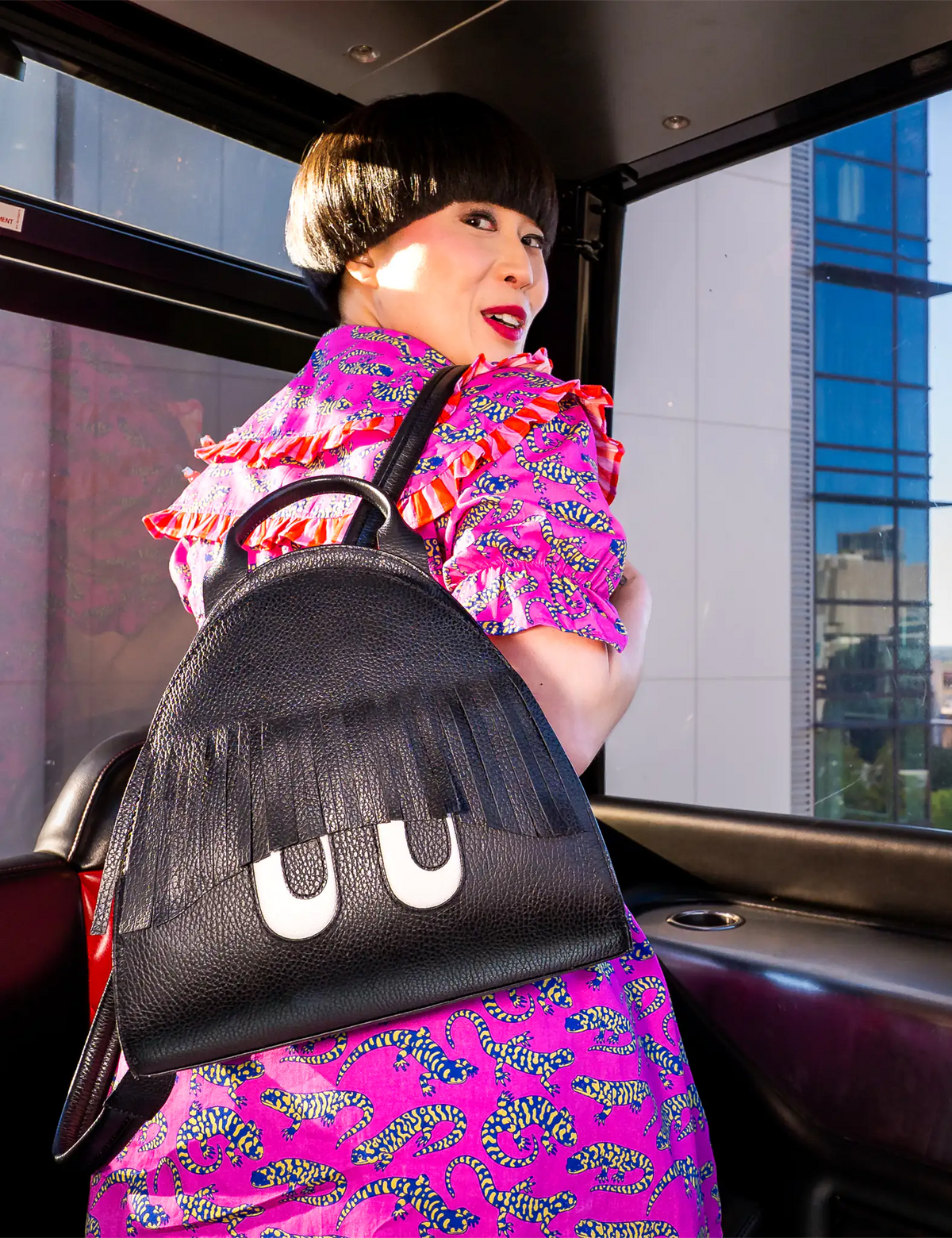 Model in a vibrant pink patterned outfit poses inside a ferris wheel, showcasing a black Min & Mon backpack featuring playful fringe bangs and engraved cartoon eyes.