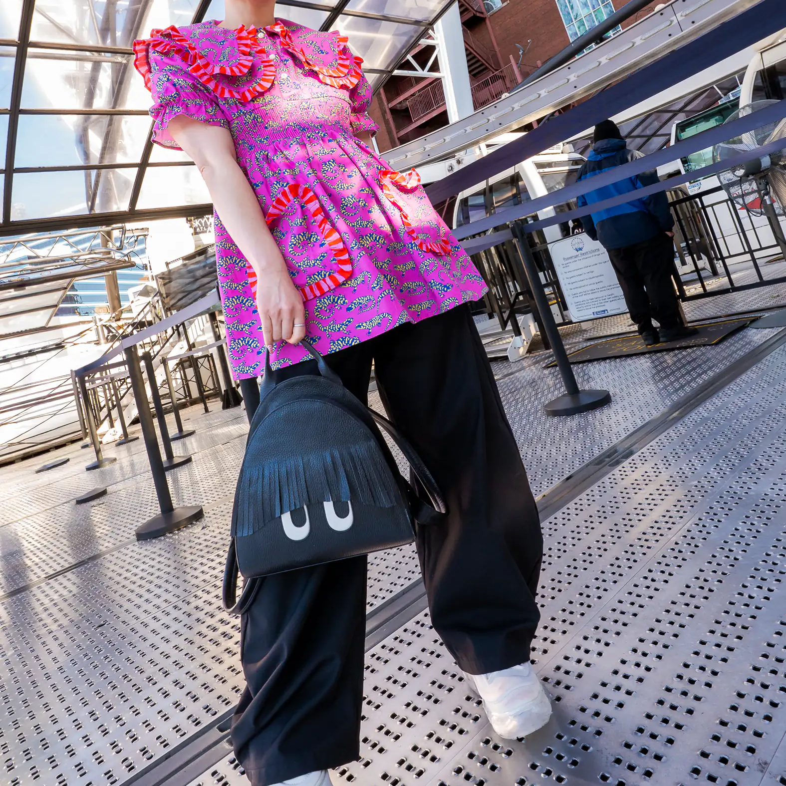 Woman in a vibrant pink patterned top, black pants, and white sneakers carries a black Min & Mon backpack with fringe bangs and playful eyes while posing on a metal walkway.