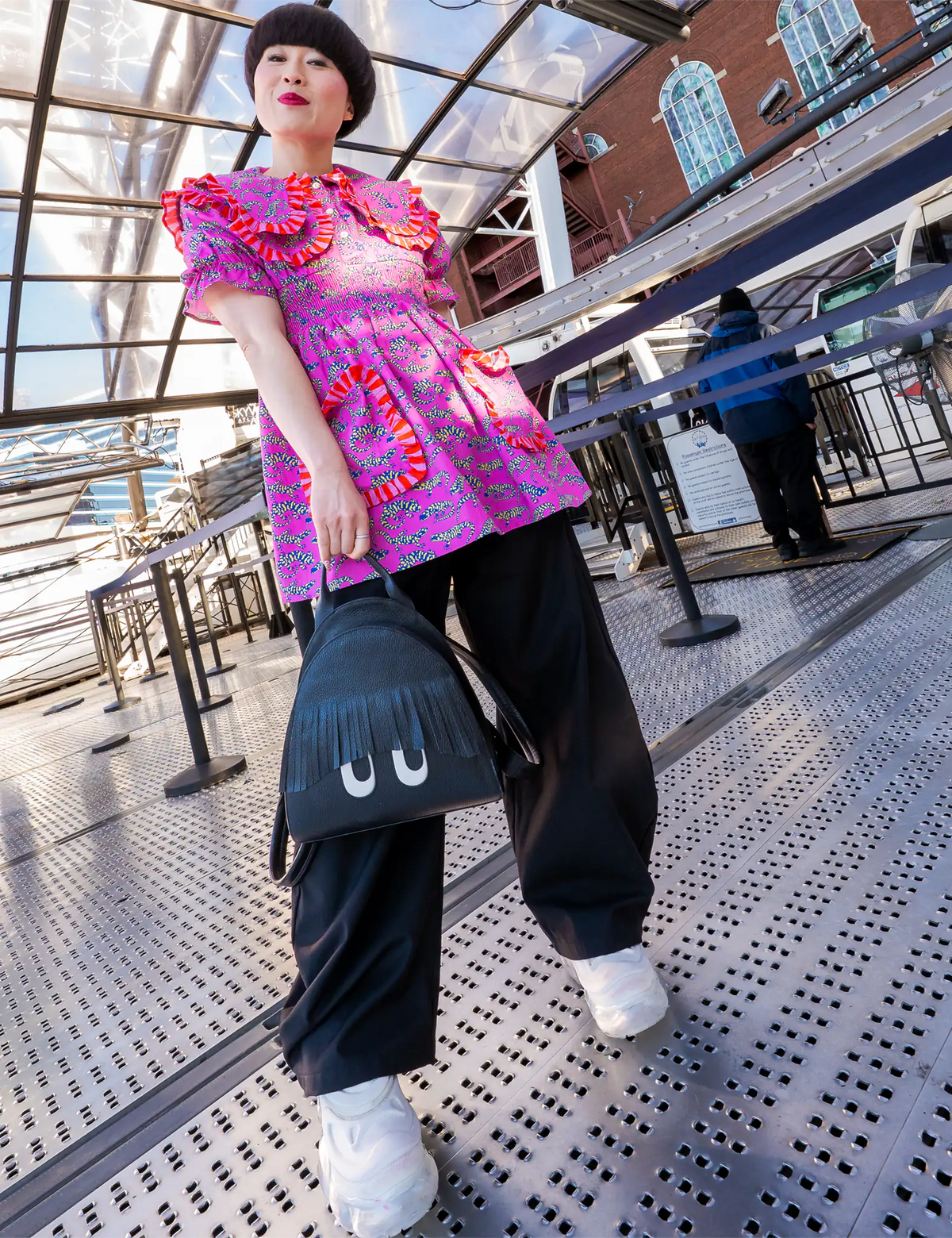 Woman in a vibrant pink patterned top, black pants, and white sneakers carries a black Min & Mon backpack with fringe bangs and playful eyes while posing on a metal walkway.