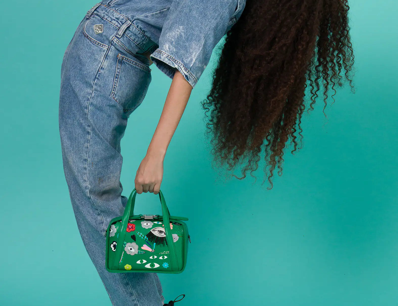Woman in denim outfit leans back, holding a green handbag with quirky designs. Her long, curly hair flows down against a teal background. Energetic mood.