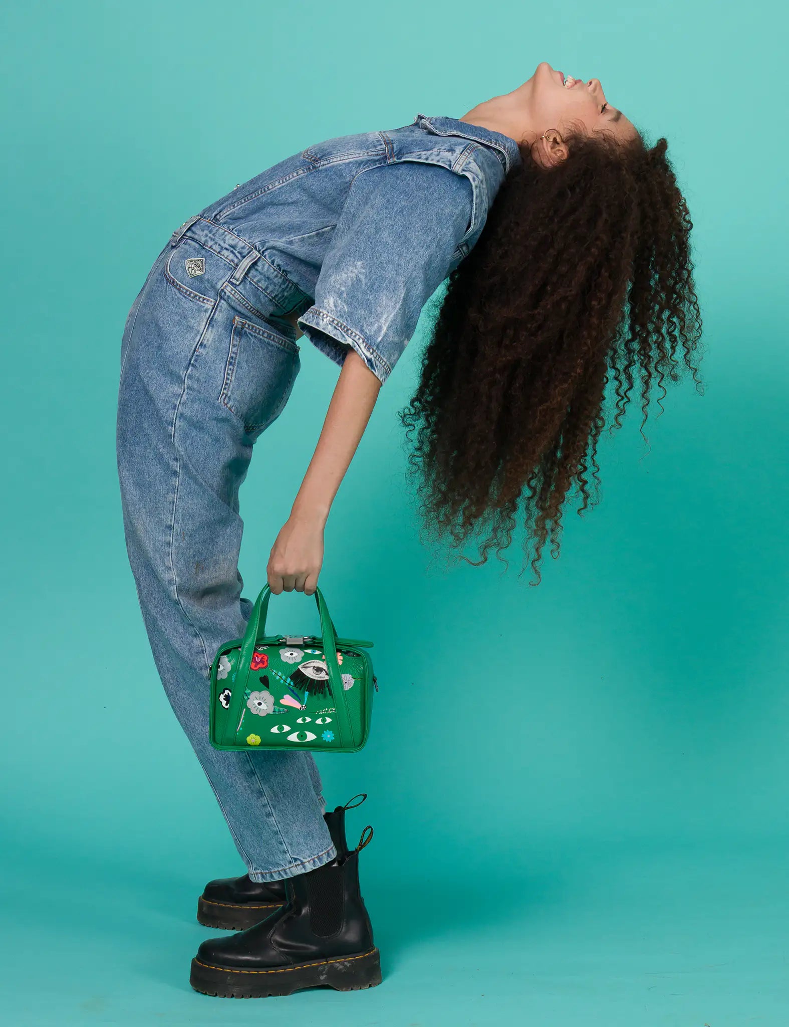 Woman in denim outfit leans back, holding a green handbag with quirky designs. Her long, curly hair flows down against a teal background. Energetic mood.