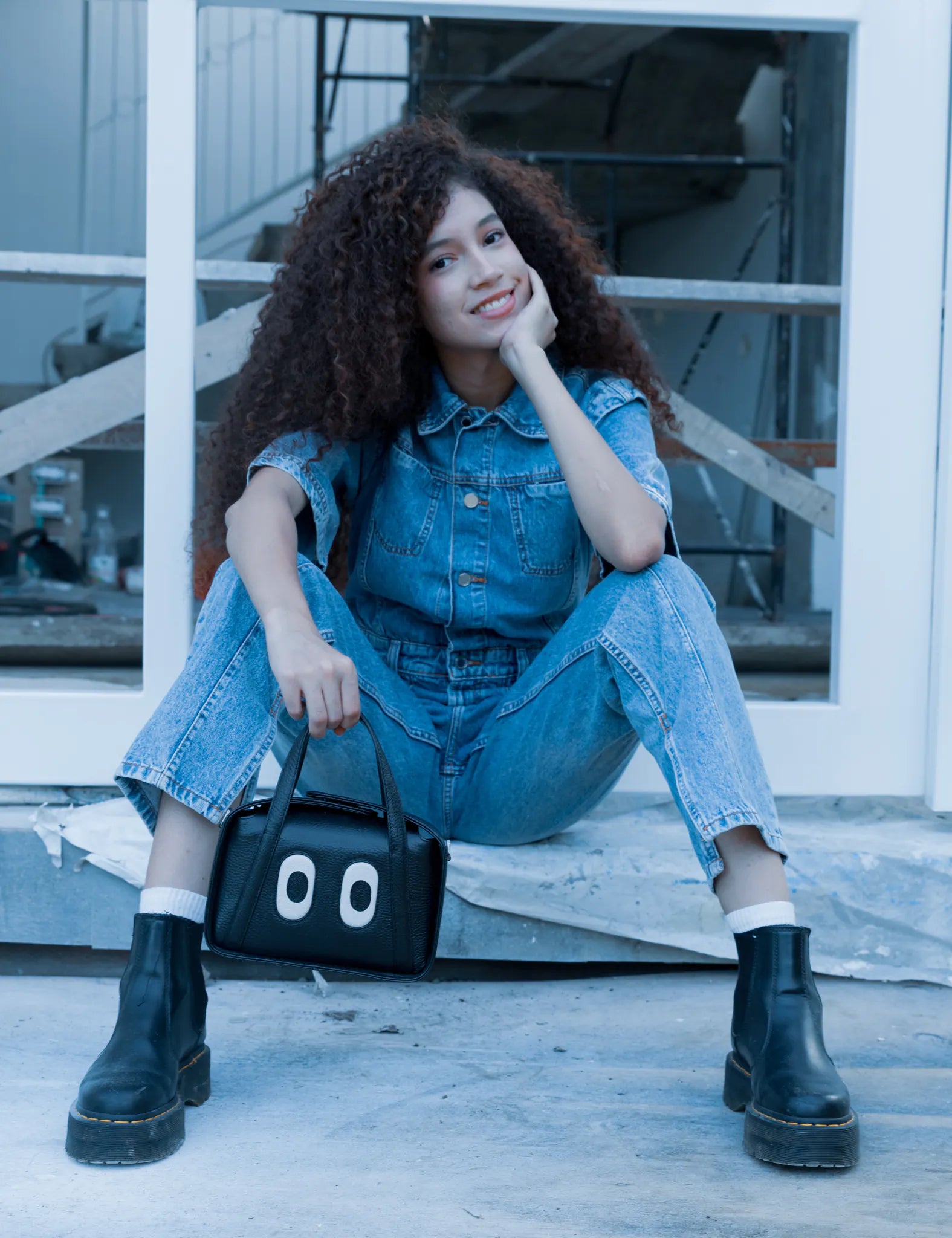 Woman with curly hair sits casually on a step in a denim jumpsuit and black boots, smiling while holding a small black handbag with playful eye motifs in an industrial setting.