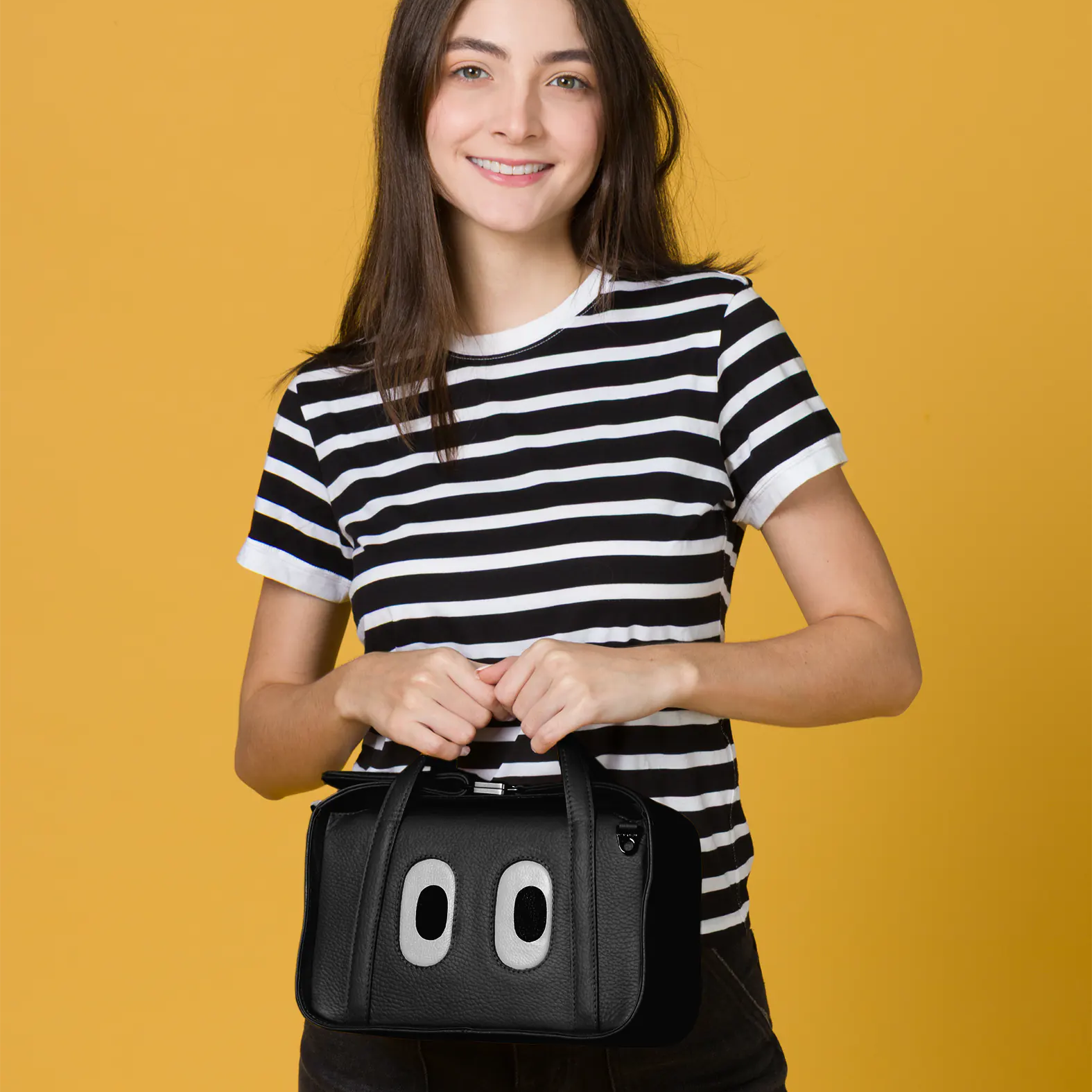 A smiling woman in a striped shirt holds the Benji Medium Crossbody Black Leather Bag from Min & Mon’s "Look Up" collection, featuring signature eye motifs, against a yellow backdrop.