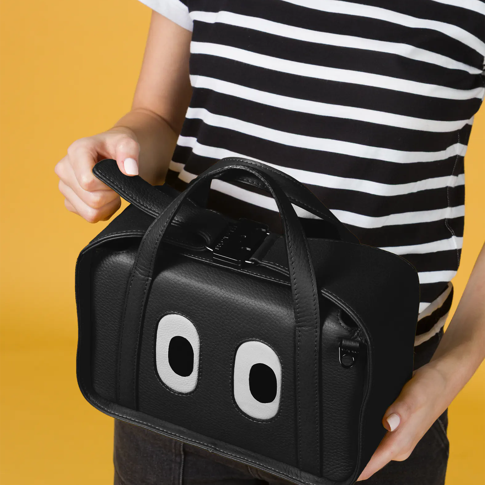 Close-up of a person holding the Benji Medium Crossbody Black Leather Bag from Min & Mon’s "Look Up" collection, featuring playful eye details and structured handles.