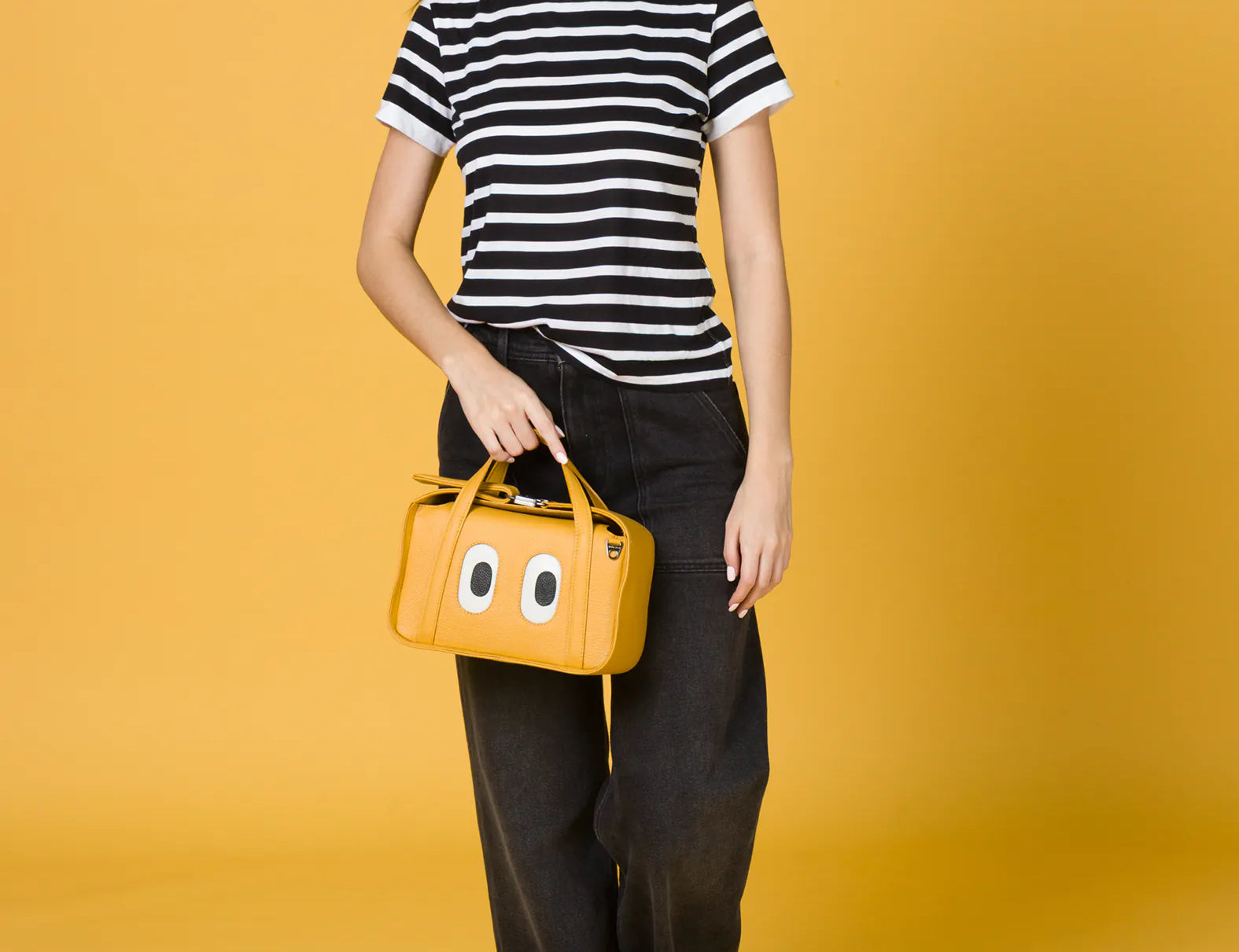 Woman walking against a yellow background, wearing a black-and-white striped t-shirt, wide-leg black jeans, and white sneakers, holding a small yellow handbag with cartoon eyes on the front.