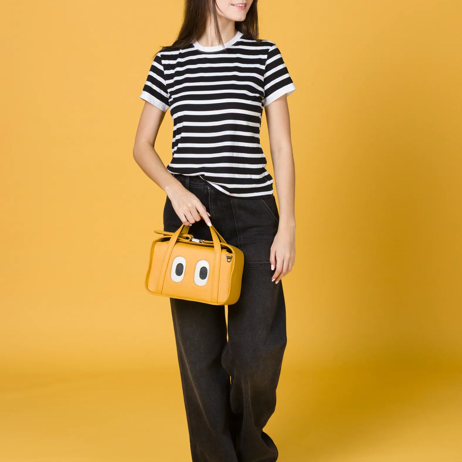 Woman walking against a yellow background, wearing a black-and-white striped t-shirt, wide-leg black jeans, and white sneakers, holding a small yellow handbag with cartoon eyes on the front.