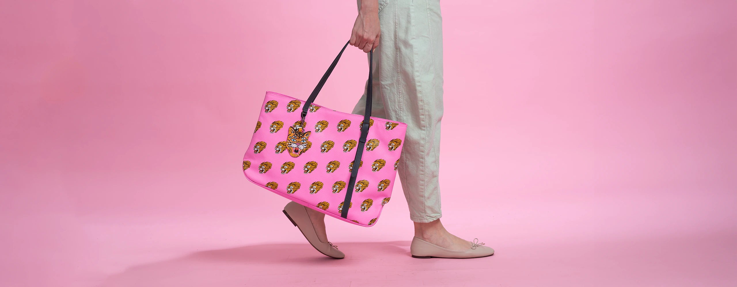 A woman wearing light beige pants and nude flats is carrying a vibrant pink tote bag adorned with multiple tiger head motifs. The bag features black handles and a tiger face charm. The background is a solid pink color, complementing the bold design of the tote bag.