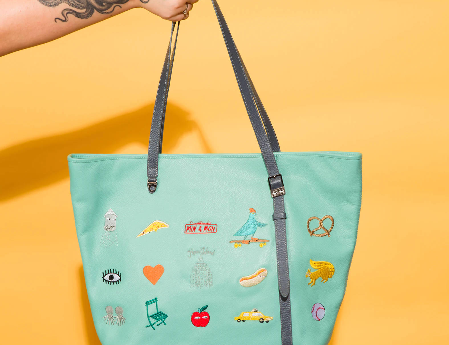 Hand with octopus tattoo holds a mint green tote bag featuring colorful NYC-themed embroidered patches like a pigeon on a skateboard, pretzel, taxi, and hot dog. Set against a yellow background.