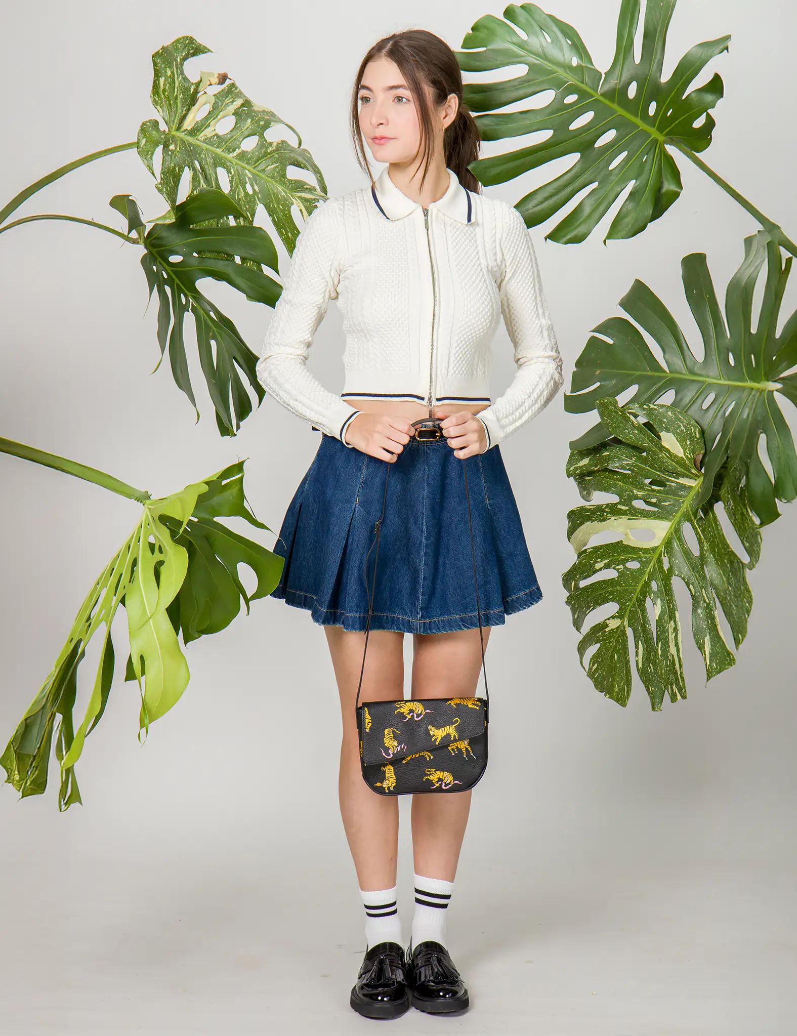 Woman in white sweater and denim skirt stands among large green leaves, holding a small black crossbody bag decorated with embroidered yellow tigers.