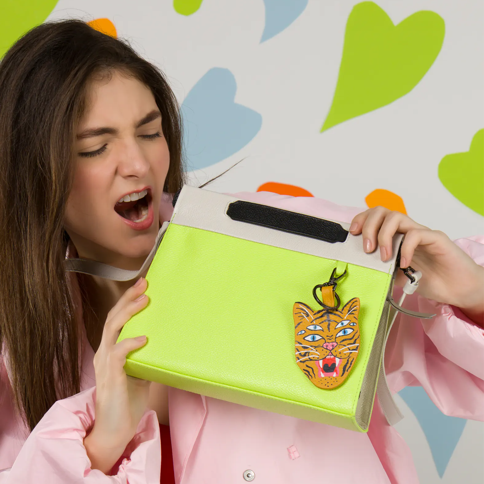 A woman in a pink jacket holds a bright lime green handbag with a black handle and a tiger face charm, set against a white wall decorated with colorful heart shapes.