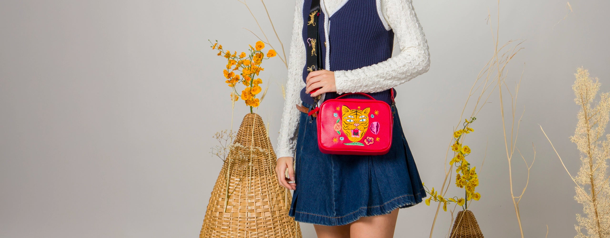 A person wearing a navy sweater vest, white textured long-sleeve top, and a denim skirt poses in a minimalist studio decorated with dried plants and wicker baskets. They are holding a vibrant pink crossbody bag featuring a bold embroidered tiger face and whimsical elements.