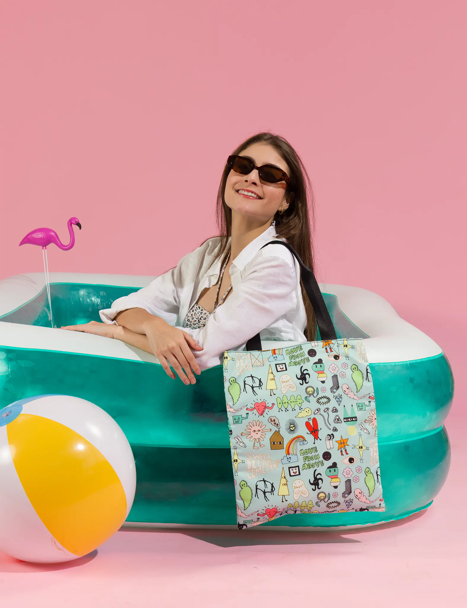 Smiling woman wearing sunglasses sits in an inflatable pool against a pink background, holding a tote bag with colorful playful illustrations; a beach ball and flamingo float add a fun vibe.
