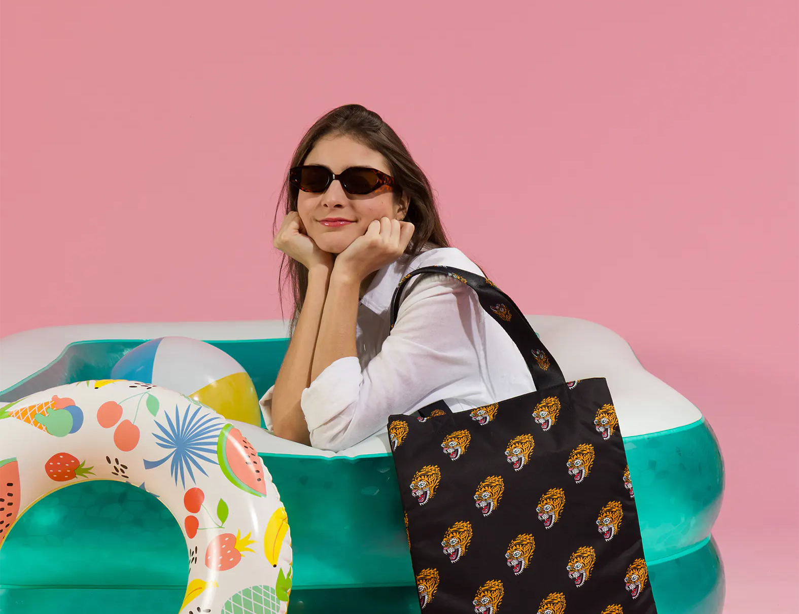 Woman in sunglasses leans on an inflatable pool against a pink background, holding a black tote bag with repeating tiger face prints; a colorful pool float adds a playful summer vibe.