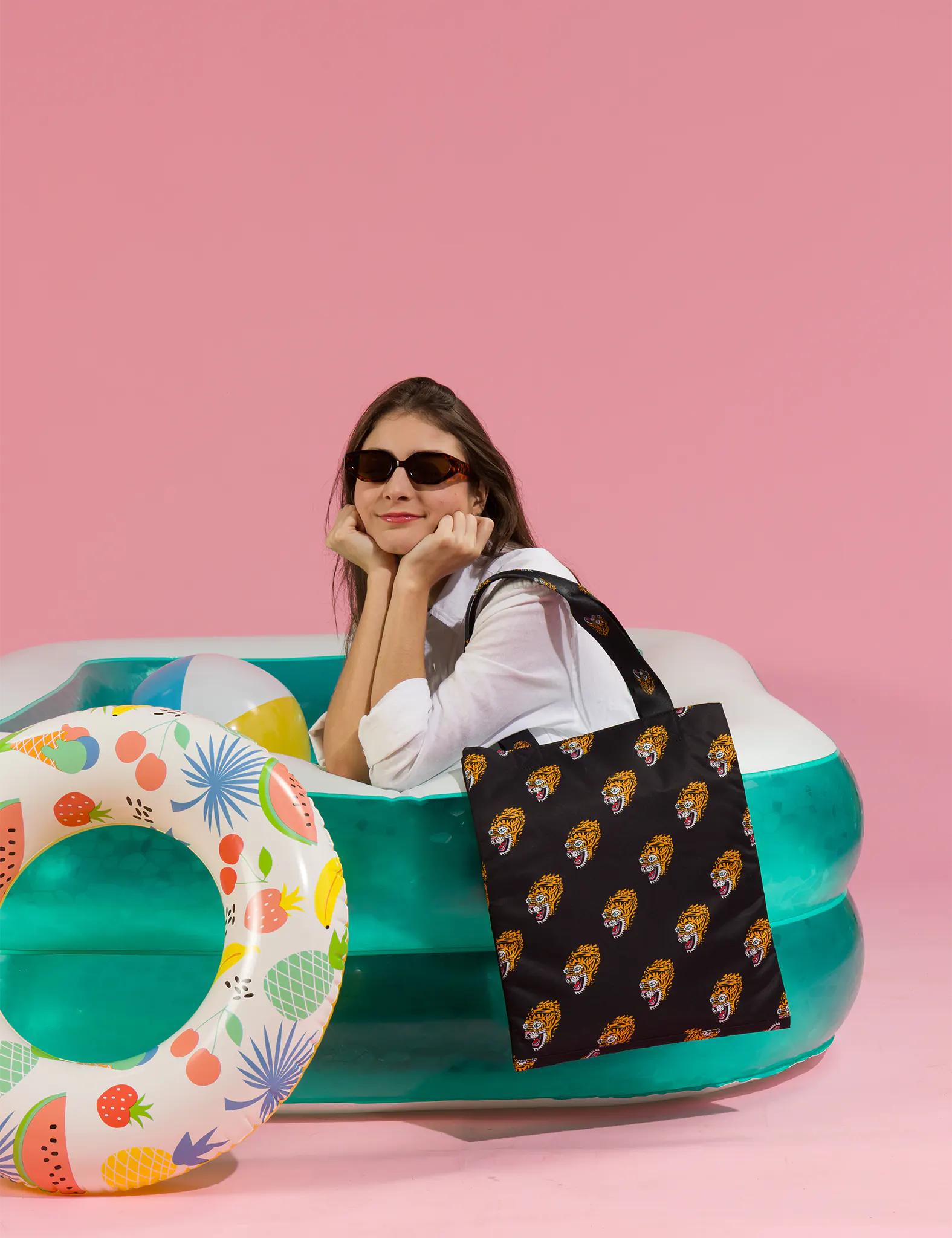 Woman in sunglasses leans on an inflatable pool against a pink background, holding a black tote bag with repeating tiger face prints; a colorful pool float adds a playful summer vibe.
