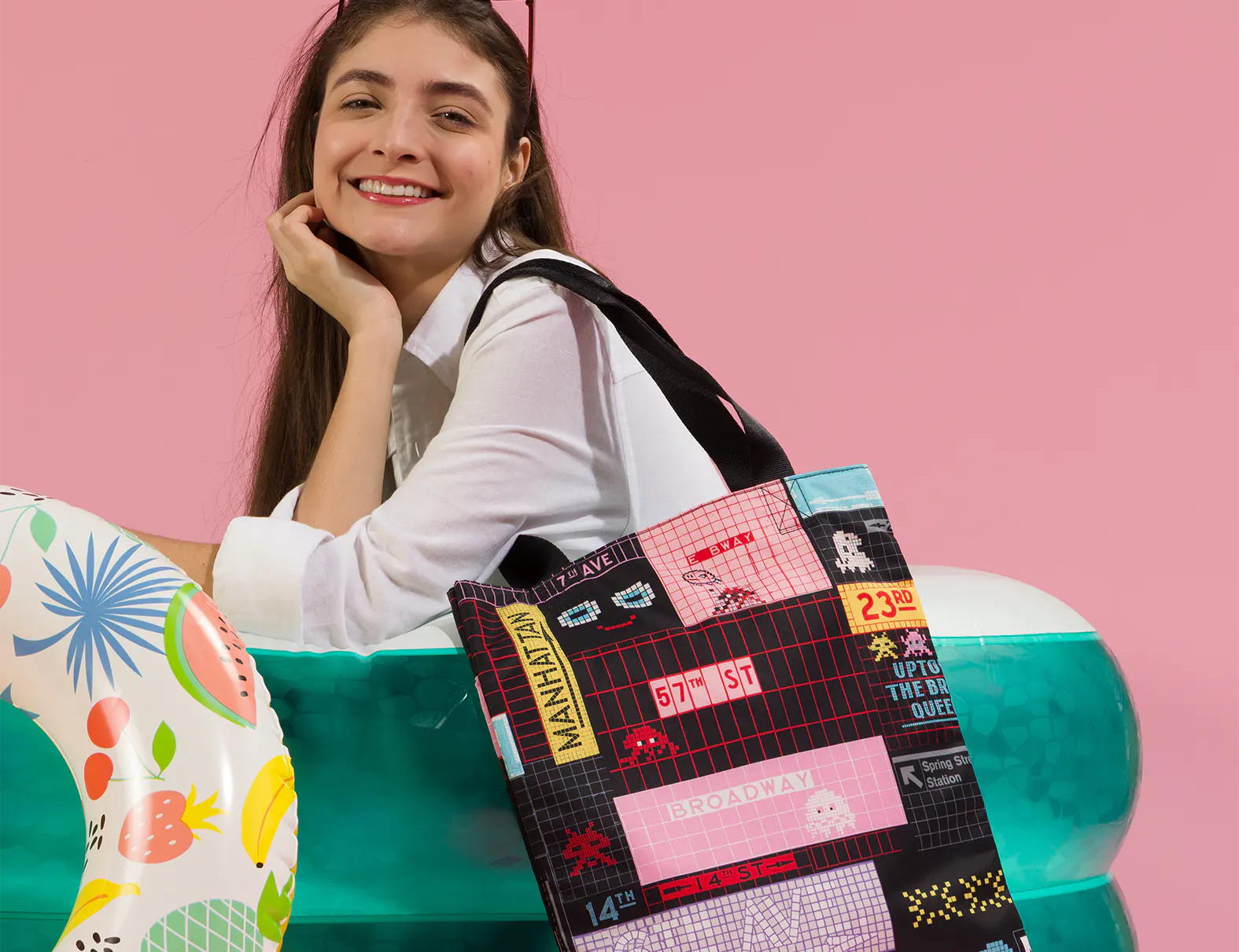 Smiling woman with sunglasses on her head sits in an inflatable pool against a pink background, holding a tote bag with colorful NYC-inspired graphic prints; a patterned pool float adds a playful touch.