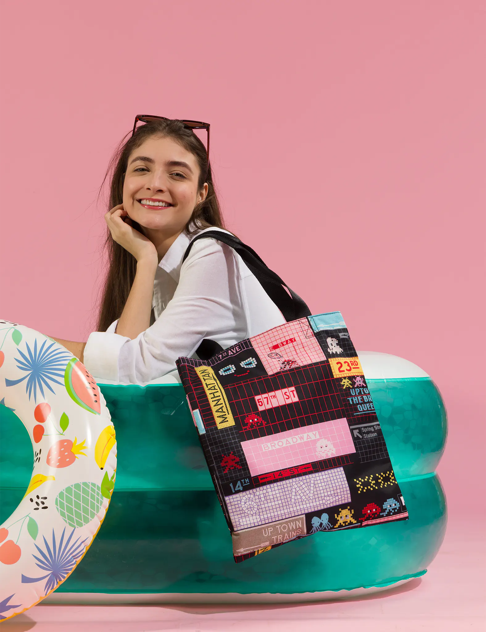 Smiling woman with sunglasses on her head sits in an inflatable pool against a pink background, holding a tote bag with colorful NYC-inspired graphic prints; a patterned pool float adds a playful touch.