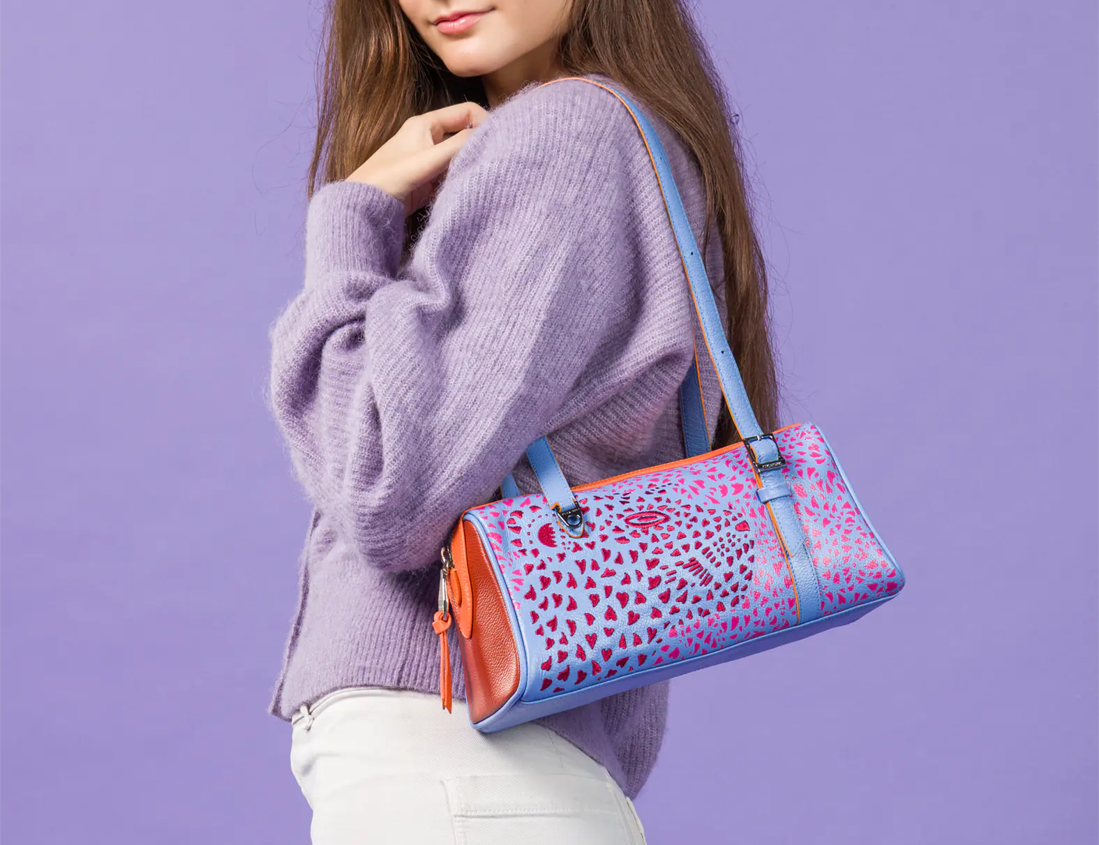 A woman wearing a lavender knit sweater and white pants carries a colorful patterned shoulder bag with blue straps against a solid lavender studio background.