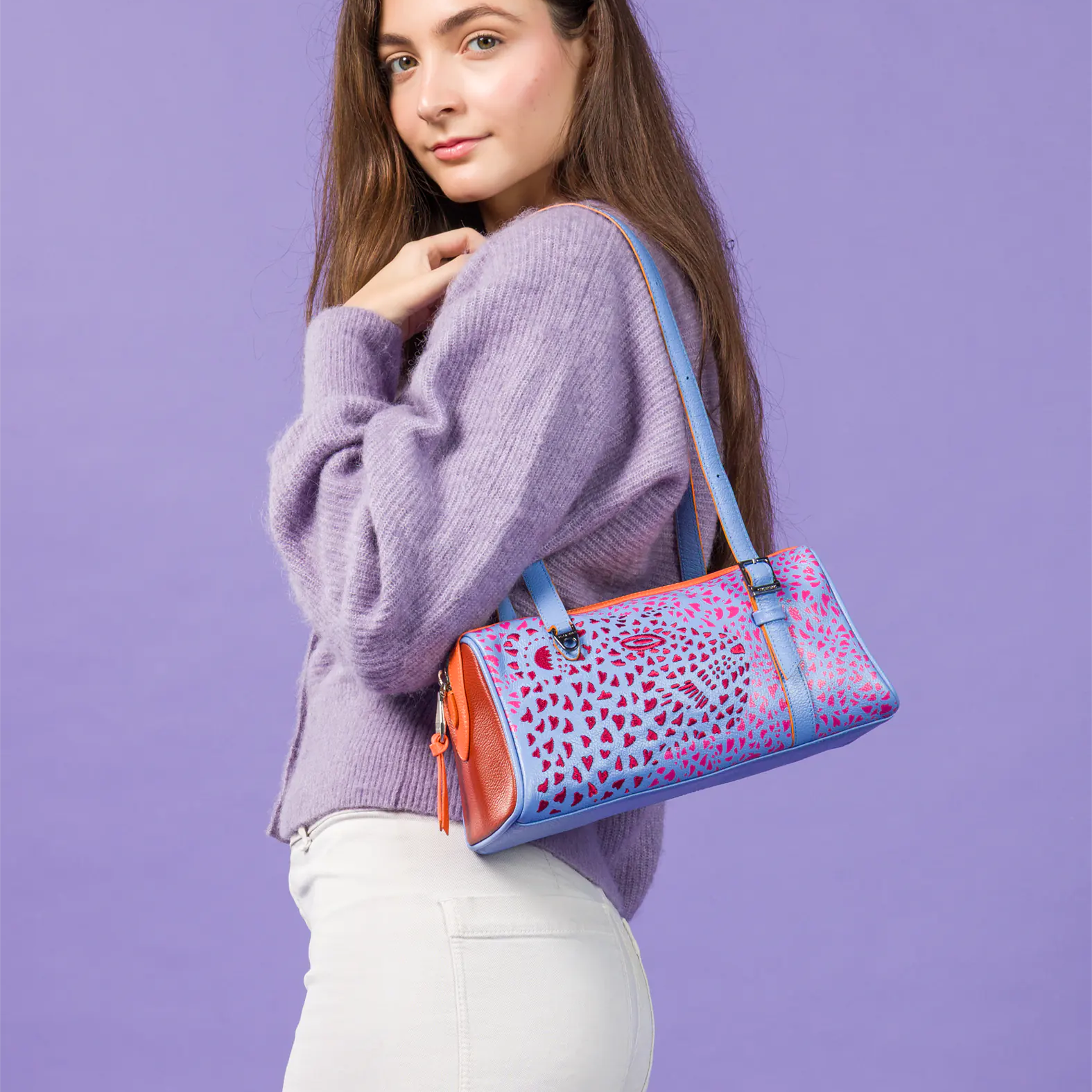 A woman wearing a lavender knit sweater and white pants carries a colorful patterned shoulder bag with blue straps against a solid lavender studio background.