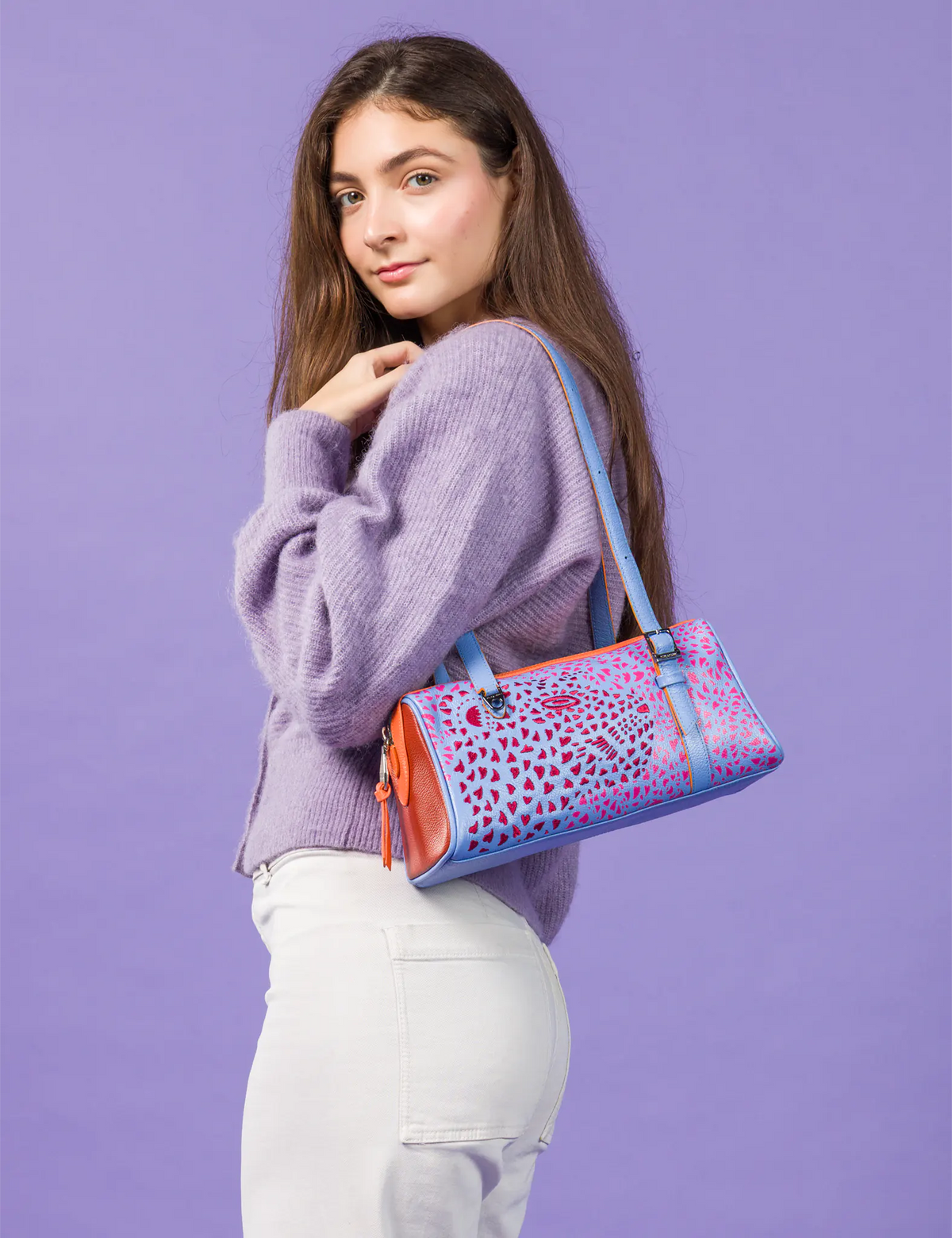 A woman wearing a lavender knit sweater and white pants carries a colorful patterned shoulder bag with blue straps against a solid lavender studio background.