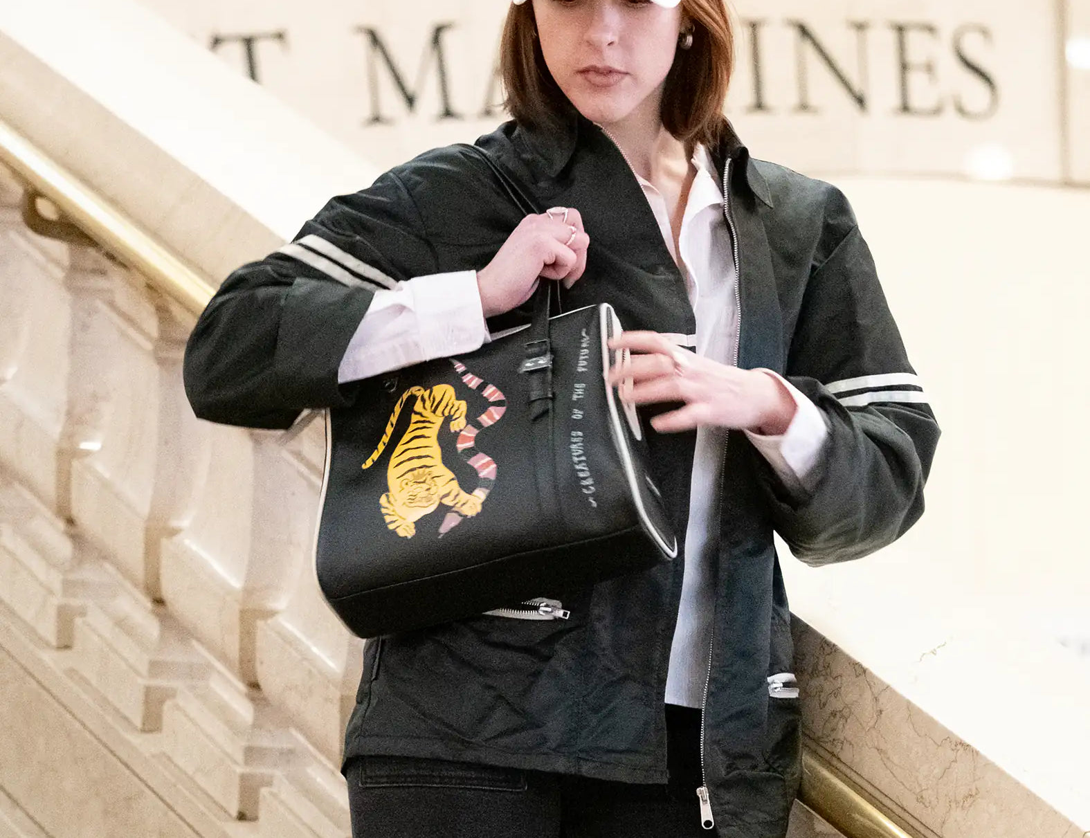 Woman in NY Yankees cap on marble staircase, holding a black handbag with a yellow tiger and pink-striped tail design. She wears a dark jacket over a white shirt and black pants.