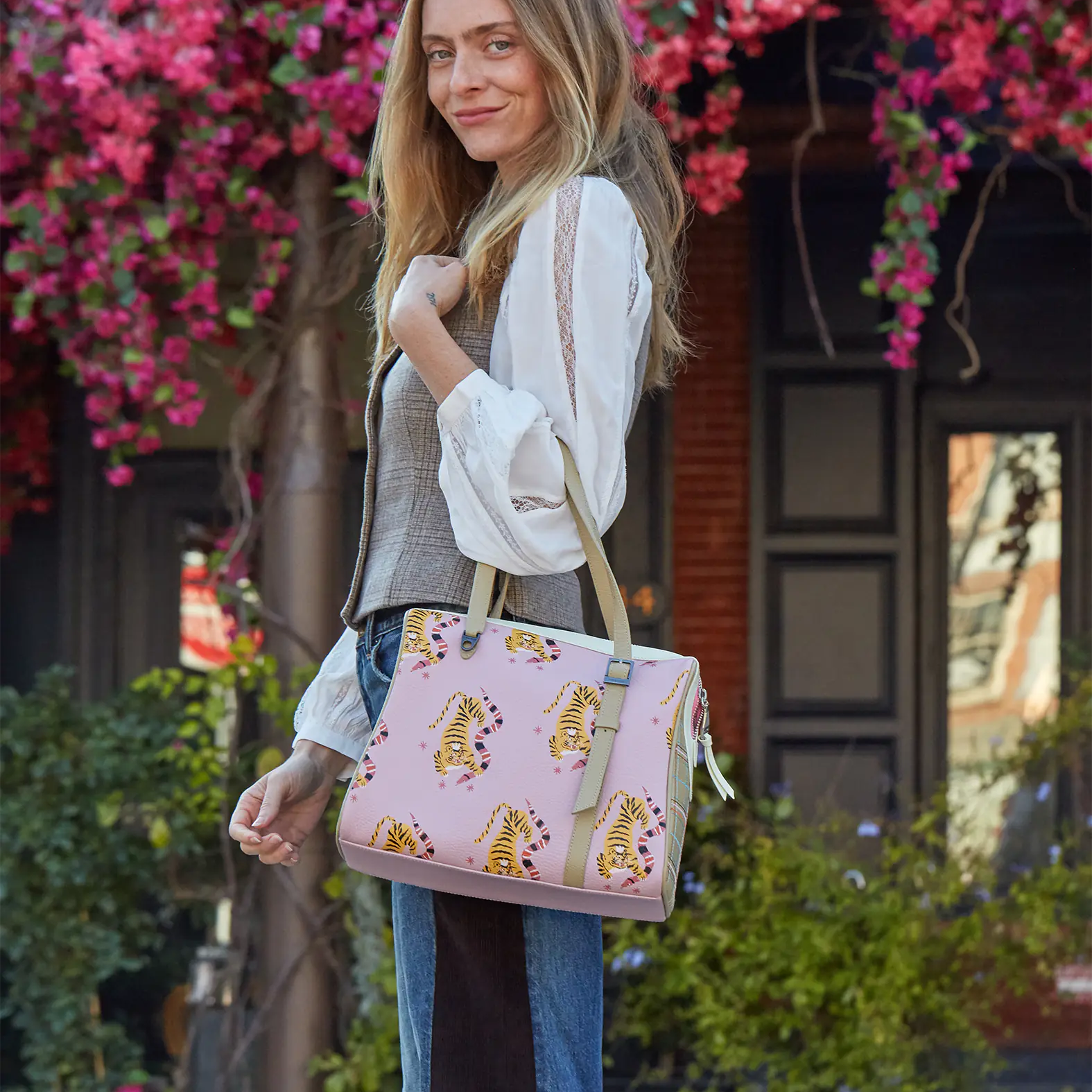 A woman standing outdoors with blooming pink flowers behind her, smiling while carrying a pink handbag decorated with yellow tiger prints.