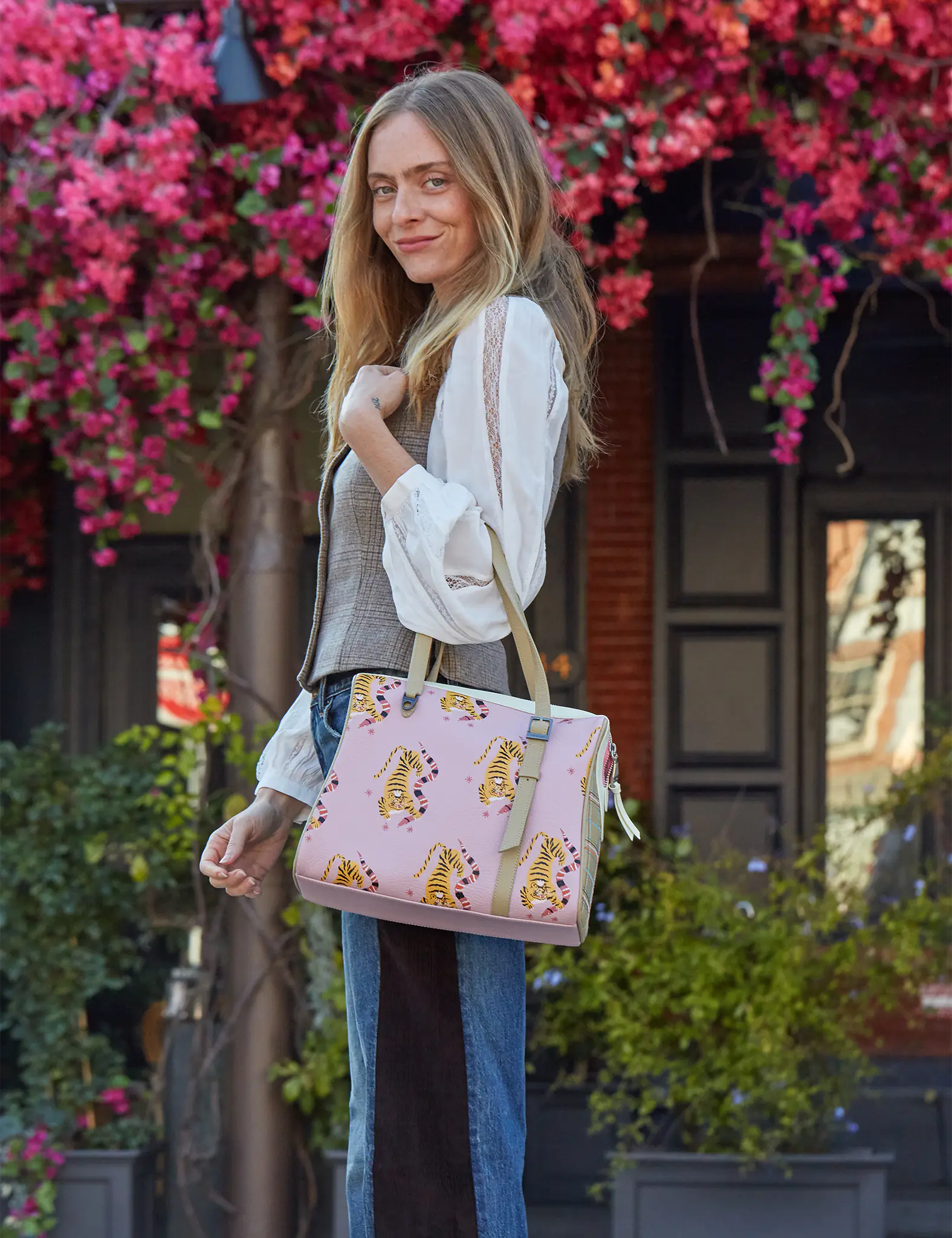 A woman standing outdoors with blooming pink flowers behind her, smiling while carrying a pink handbag decorated with yellow tiger prints.