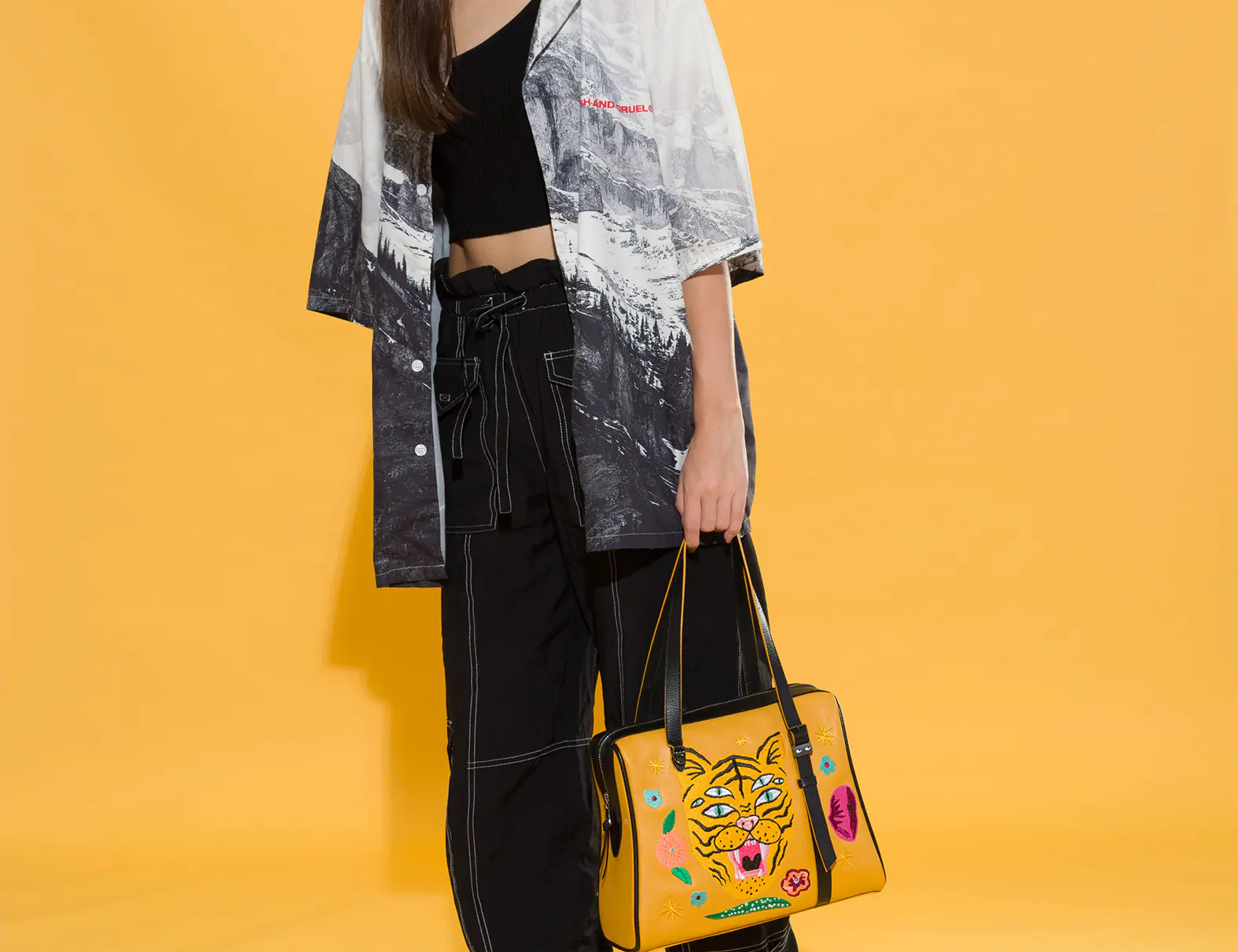 Woman in black cargo pants, platform boots, and a monochrome printed shirt holds a mustard yellow handbag embroidered with a three-eyed tiger and colorful flowers. Yellow backdrop, edgy style.