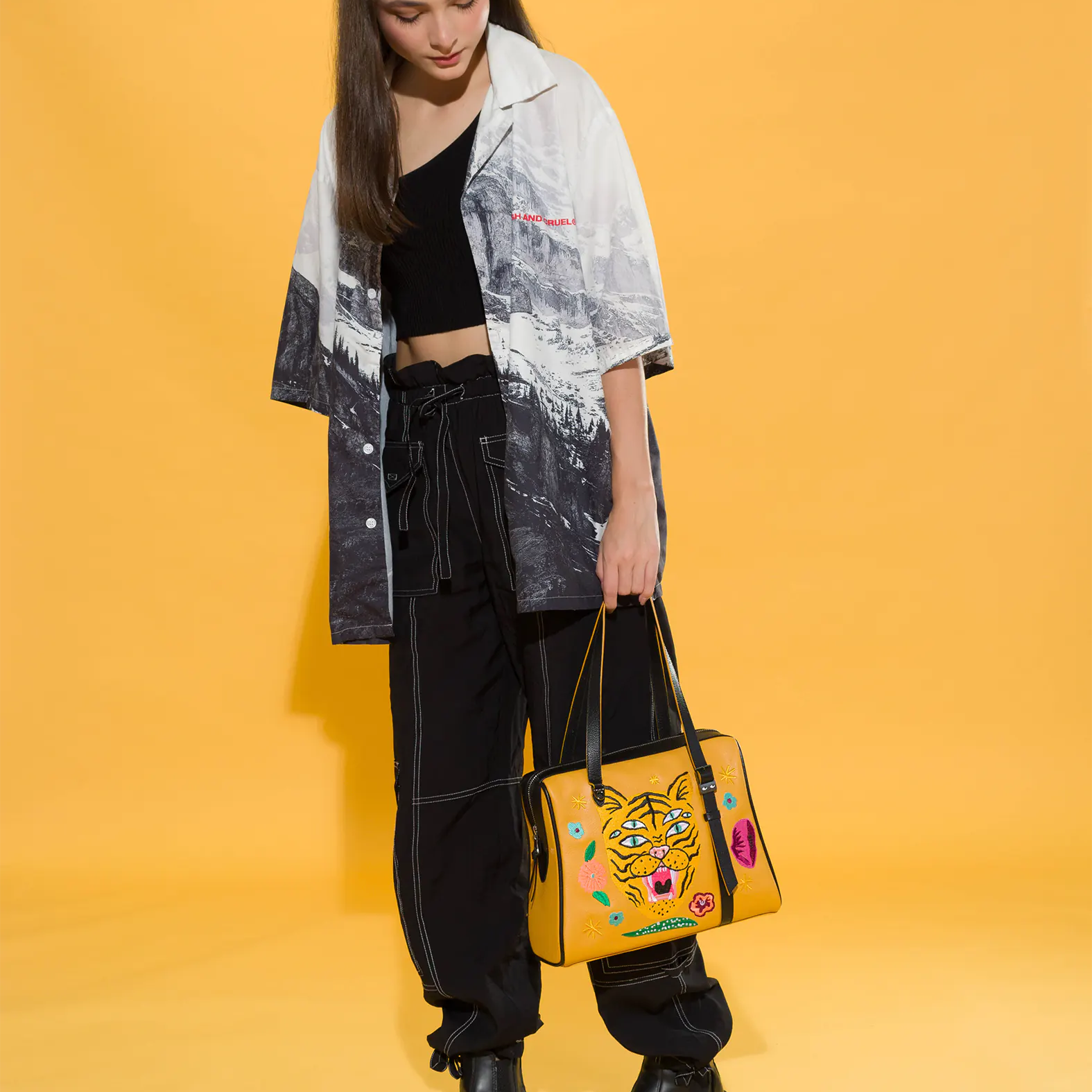 Woman in black cargo pants, platform boots, and a monochrome printed shirt holds a mustard yellow handbag embroidered with a three-eyed tiger and colorful flowers. Yellow backdrop, edgy style.