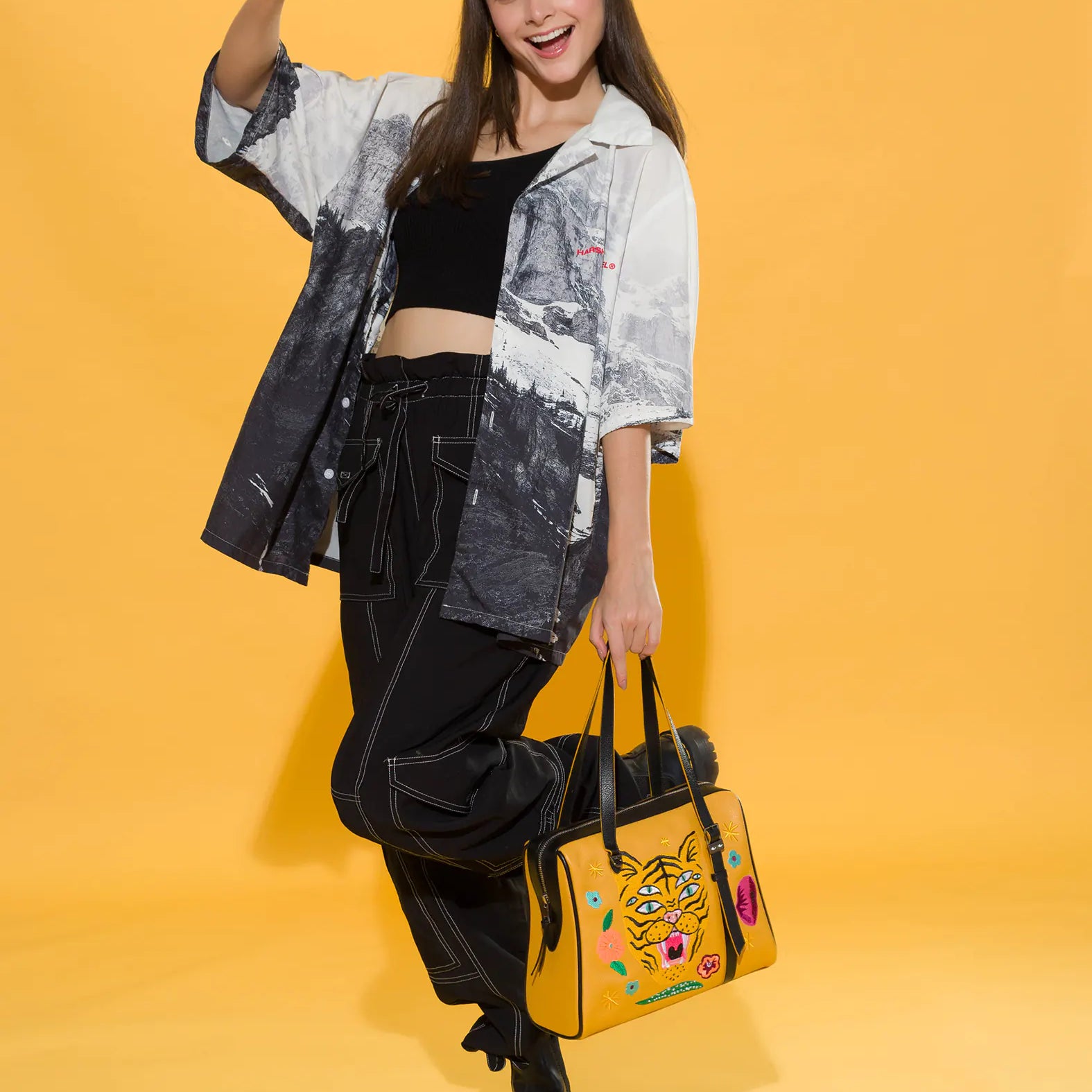 Smiling woman gives a thumbs-up while posing in black cargo pants, a cropped top, and printed shirt. She holds a mustard yellow handbag with a three-eyed tiger and floral embroidery. Yellow backdrop.
