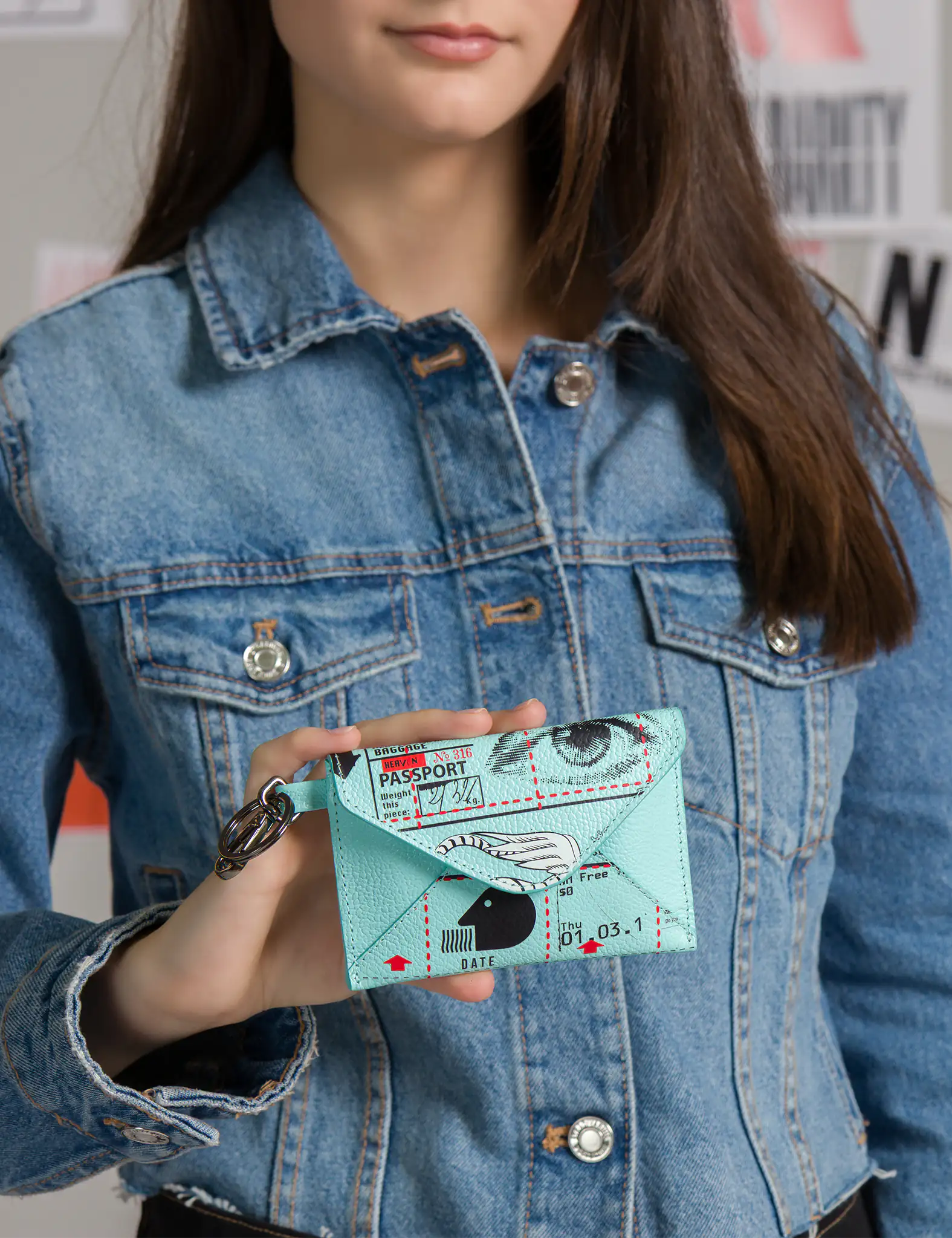 Woman in a denim jacket holds a mint-green envelope wallet with passport-stamp collage, eye and wing graphics, and a metal keyring clip; postered wall softly blurred in the background.