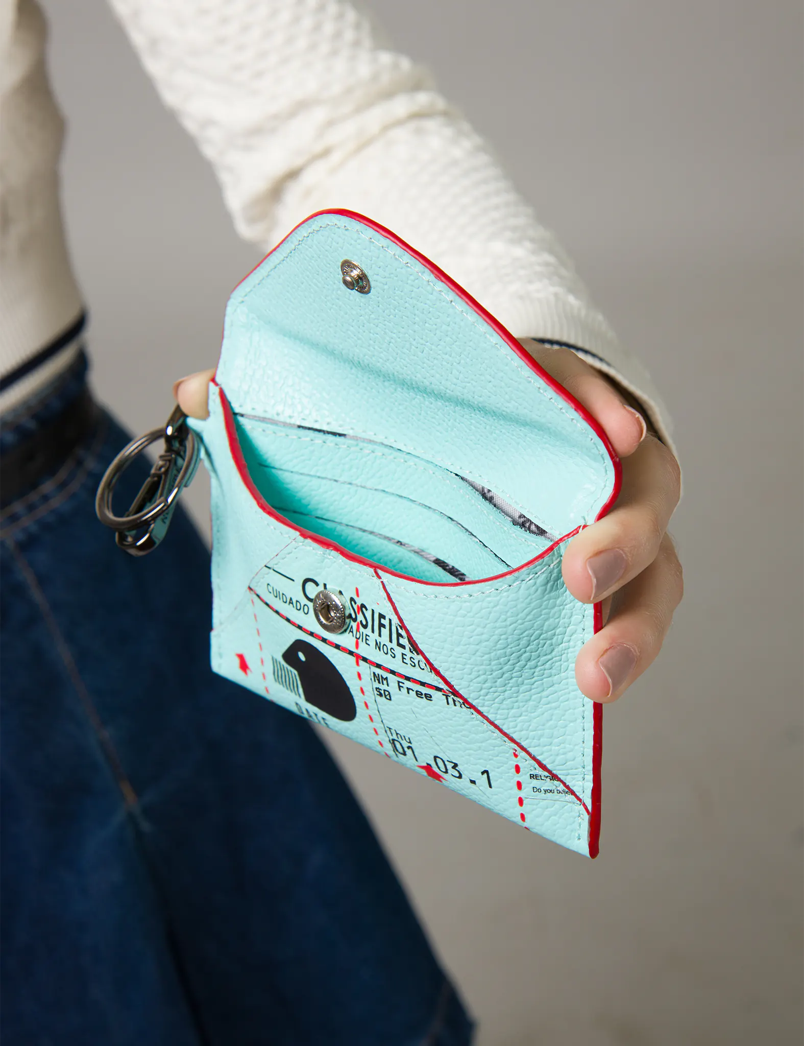 Close-up of an open turquoise leather card holder with red trim, featuring interior card slots, snap-button closure, and a metal keyring attachment on the side.
