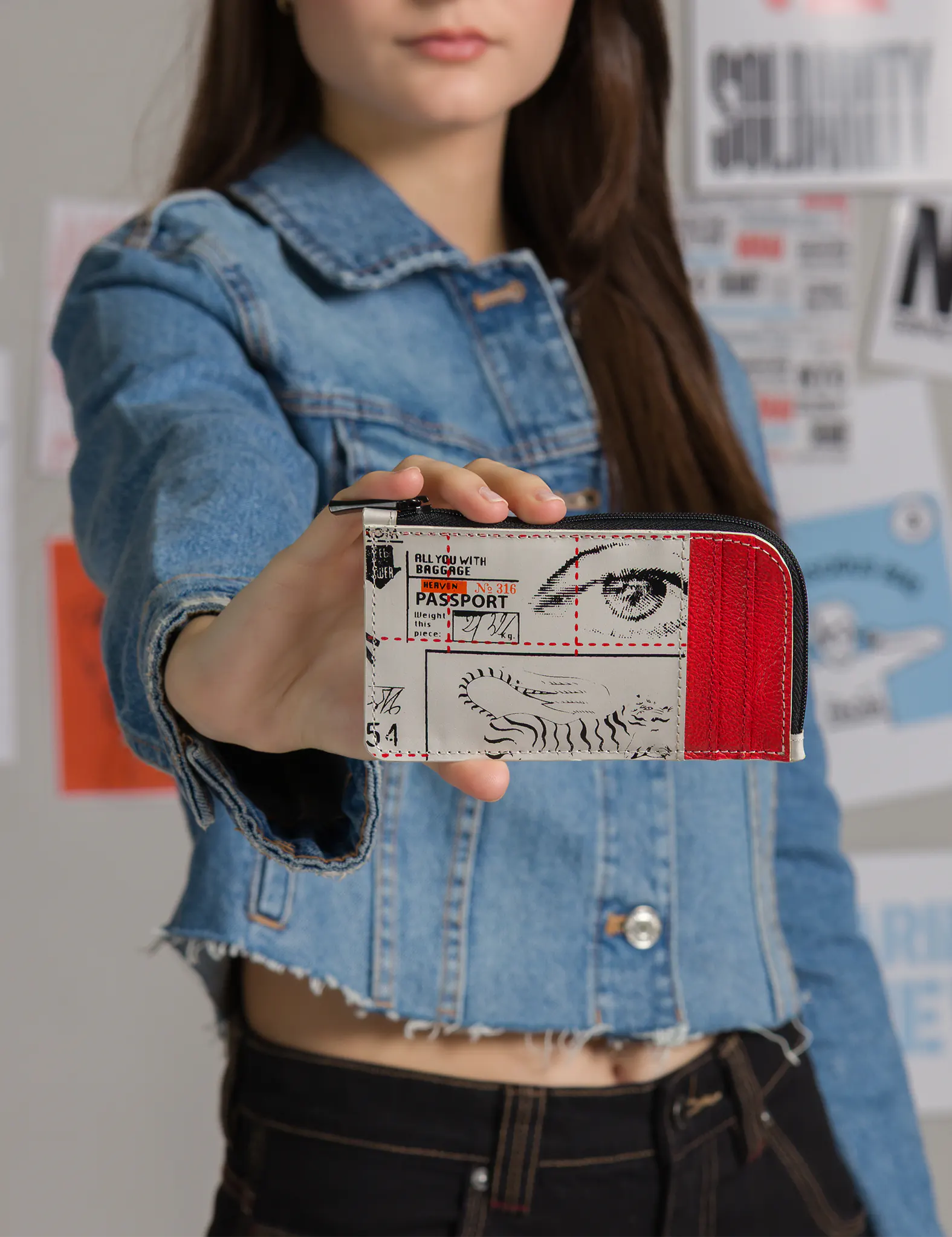 Woman in a denim jacket holds a small zip wallet toward the camera; the cream-and-red cardholder shows passport-style graphics with an illustrated eye and tiger stripes, against a poster-covered wall.