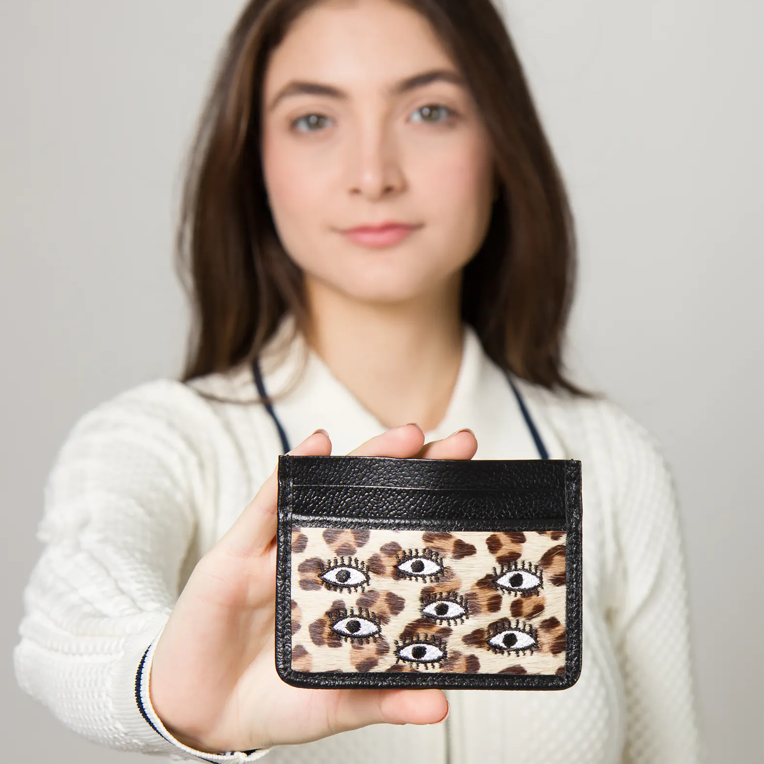 Woman holds a card holder with black leather trim and a leopard print design featuring multiple embroidered eye motifs.