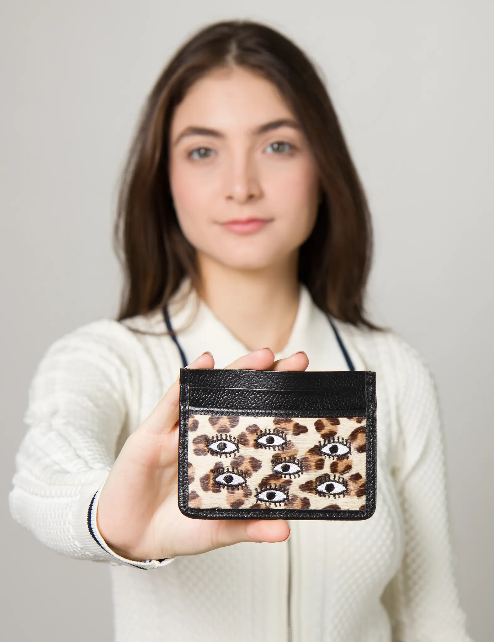 Woman holds a card holder with black leather trim and a leopard print design featuring multiple embroidered eye motifs.