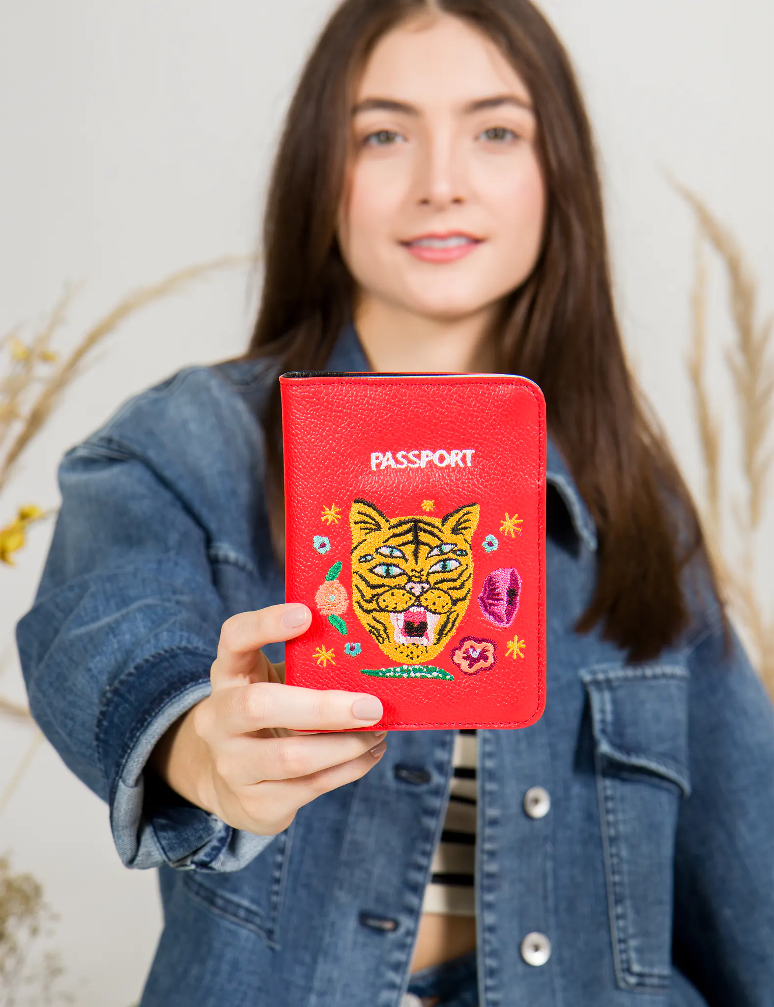 Woman in a denim jacket holds a red passport holder featuring an embroidered six-eyed tiger face surrounded by colorful floral and abstract motifs.