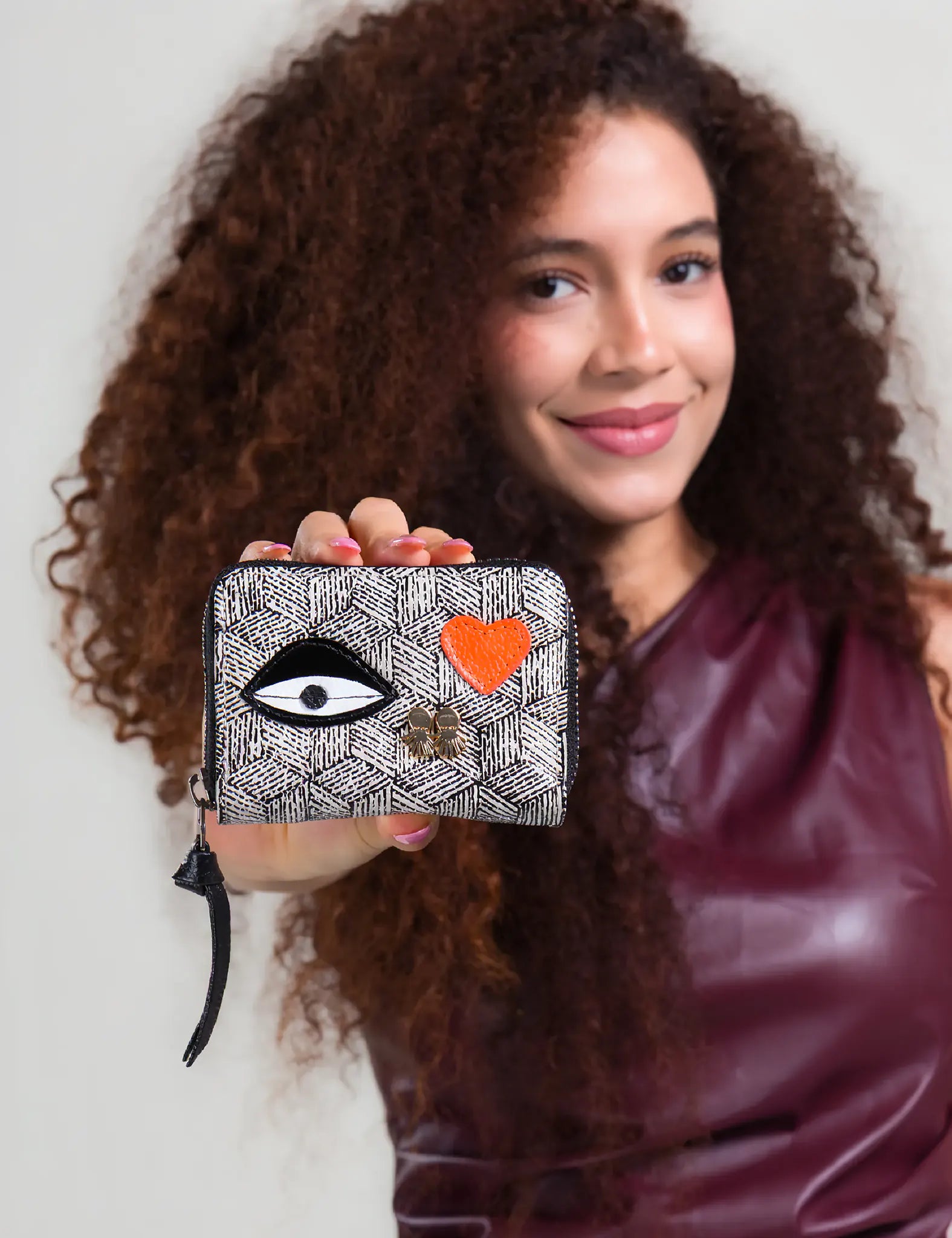 Smiling woman with curly hair holds a small zip-around wallet featuring a black-and-white geometric pattern, a large eye motif, and an orange heart detail.