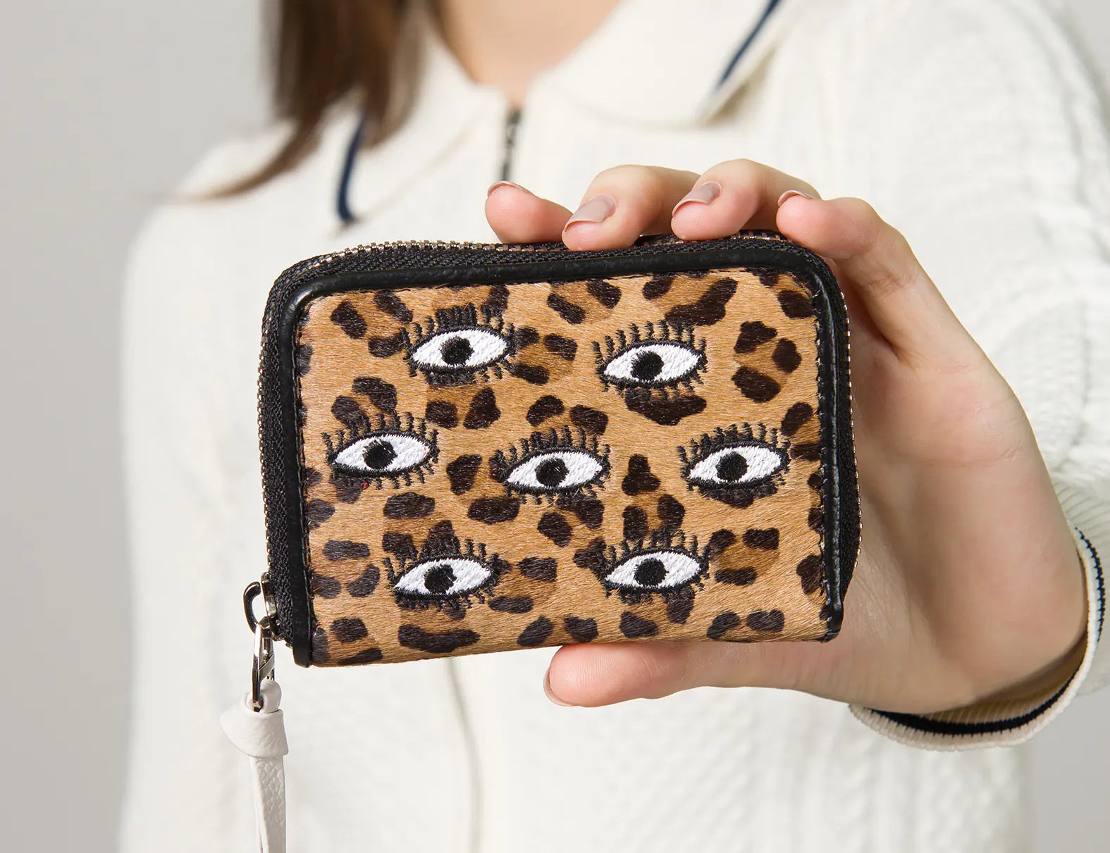 Close-up of a woman holding a leopard print zippered wallet with embroidered eye motifs, featuring a white leather zipper pull.