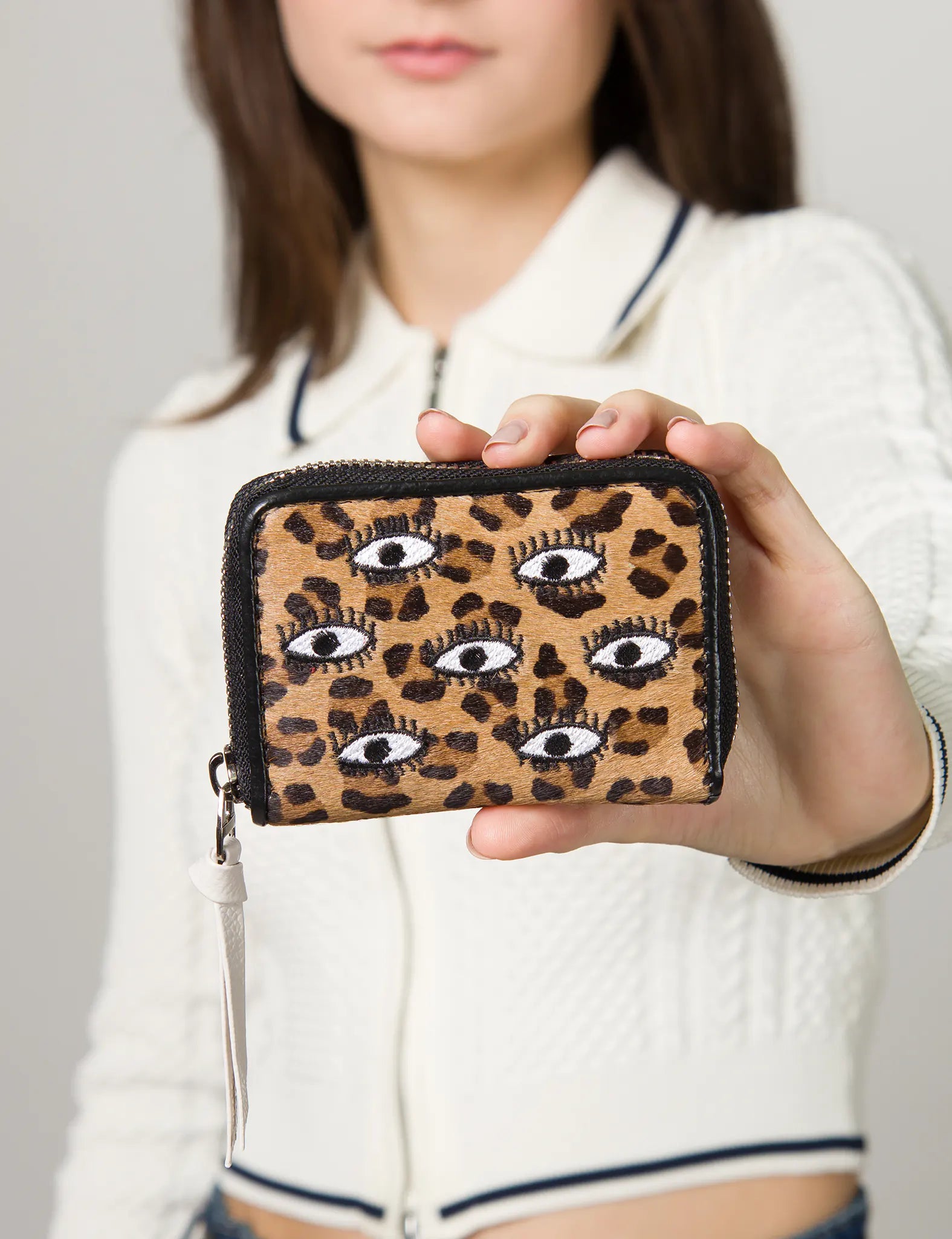 Close-up of a woman holding a leopard print zippered wallet with embroidered eye motifs, featuring a white leather zipper pull.