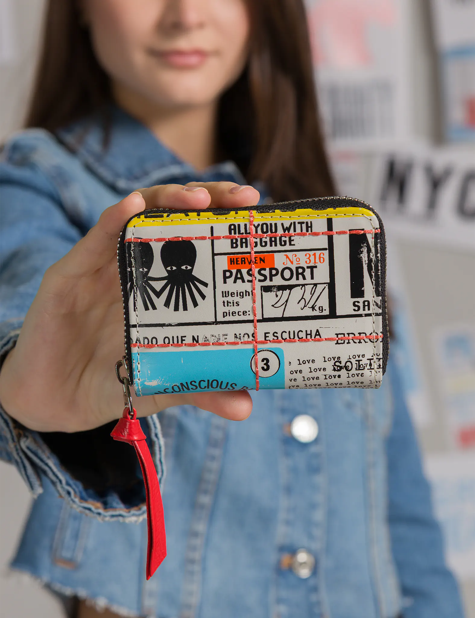 Woman in a denim jacket holds a small zip wallet toward the camera; multicolor passport-style collage with yellow top edge, aqua patch, red zipper pull, and octopus icon; posters blurred behind.