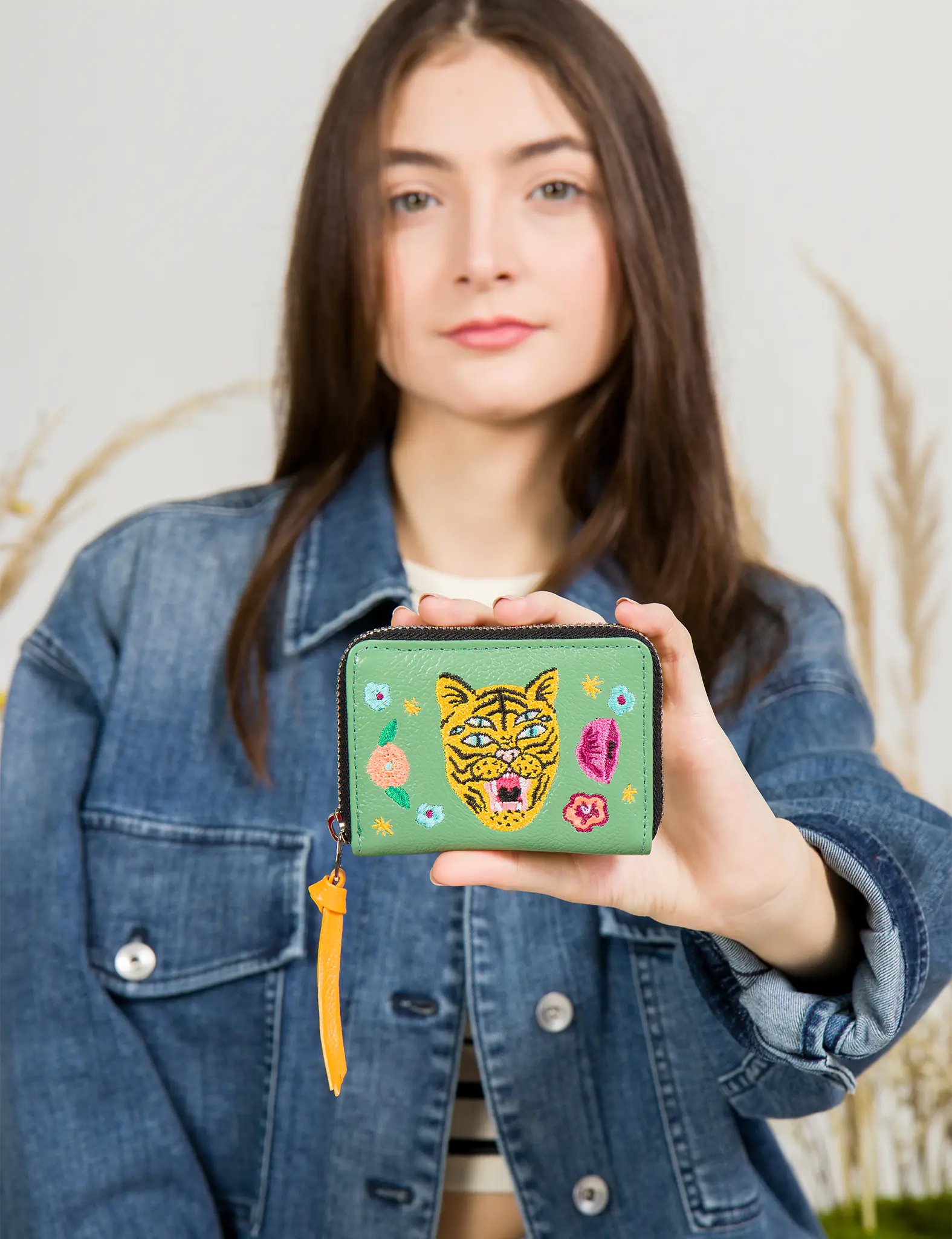 Woman in a denim jacket holds a green zip-around wallet featuring a colorful embroidered tiger face and floral motifs, with a yellow leather zipper pull.