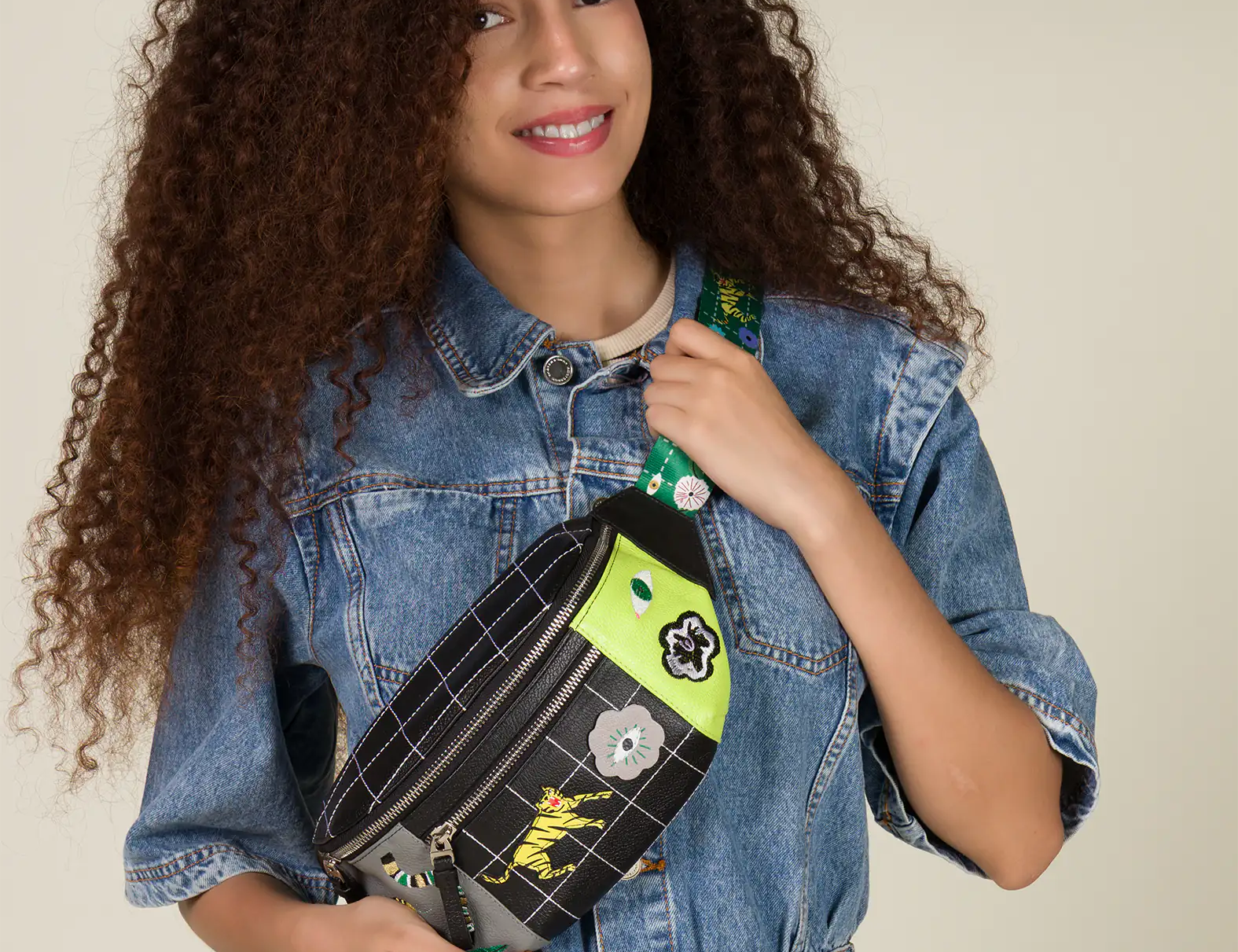 A woman with curly hair smiles warmly, wearing a denim jacket while holding a trendy Fanny Pack with a green and black pattern and flower patches.