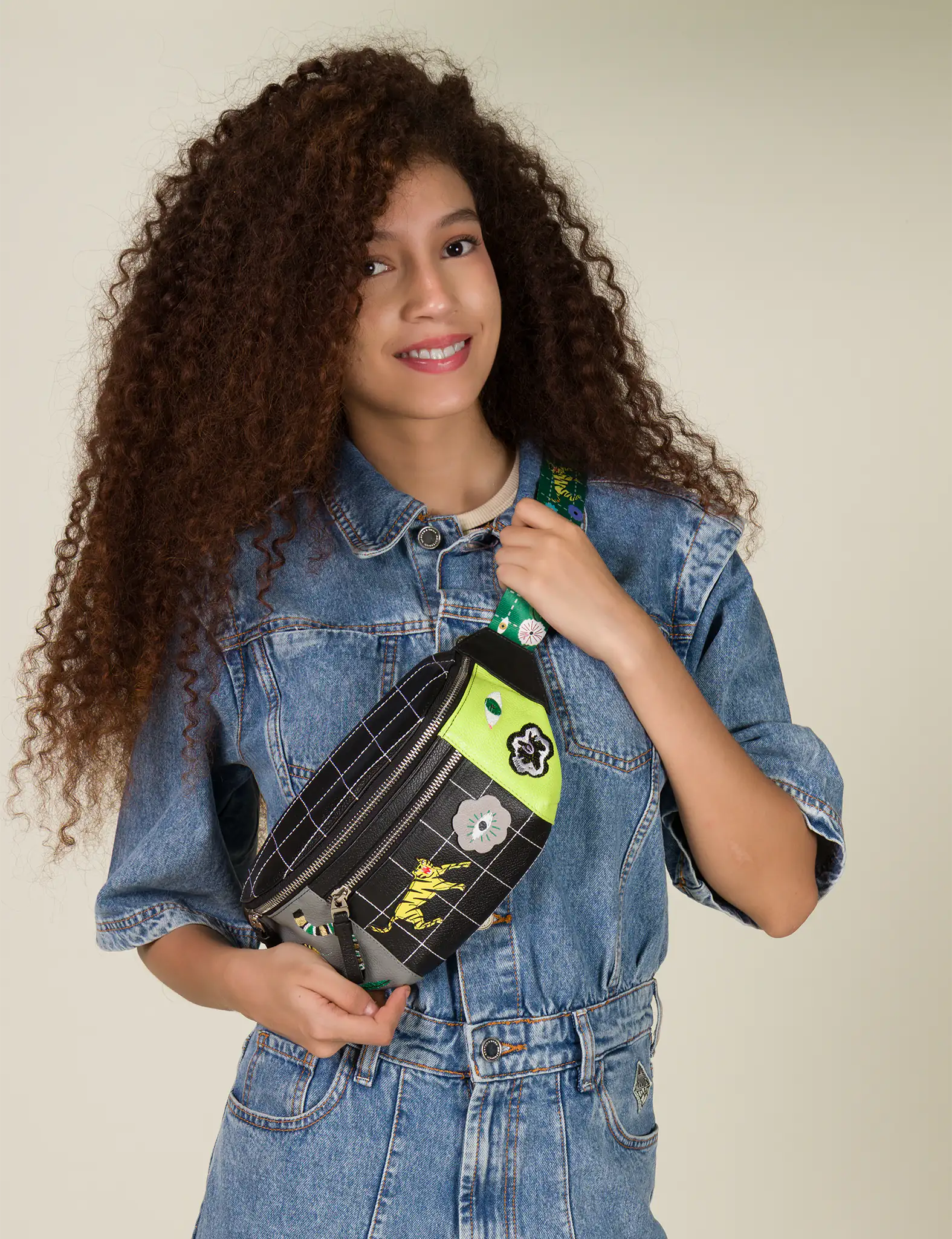 A woman with curly hair smiles warmly, wearing a denim jacket while holding a trendy Fanny Pack with a green and black pattern and flower patches.