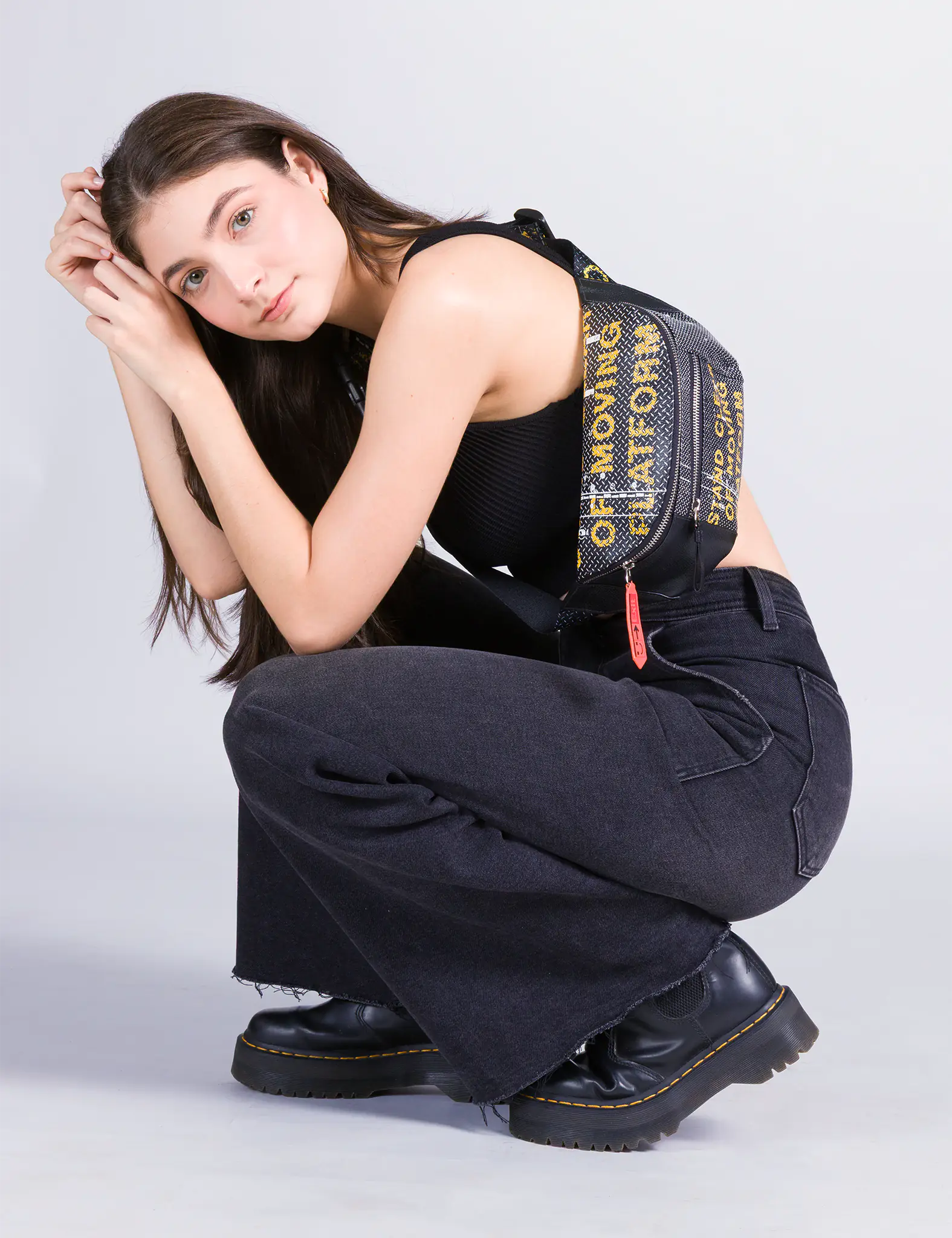 A young woman in a black outfit squats down while wearing a Min & Mon Fanny Pack with yellow text on a textured black background.