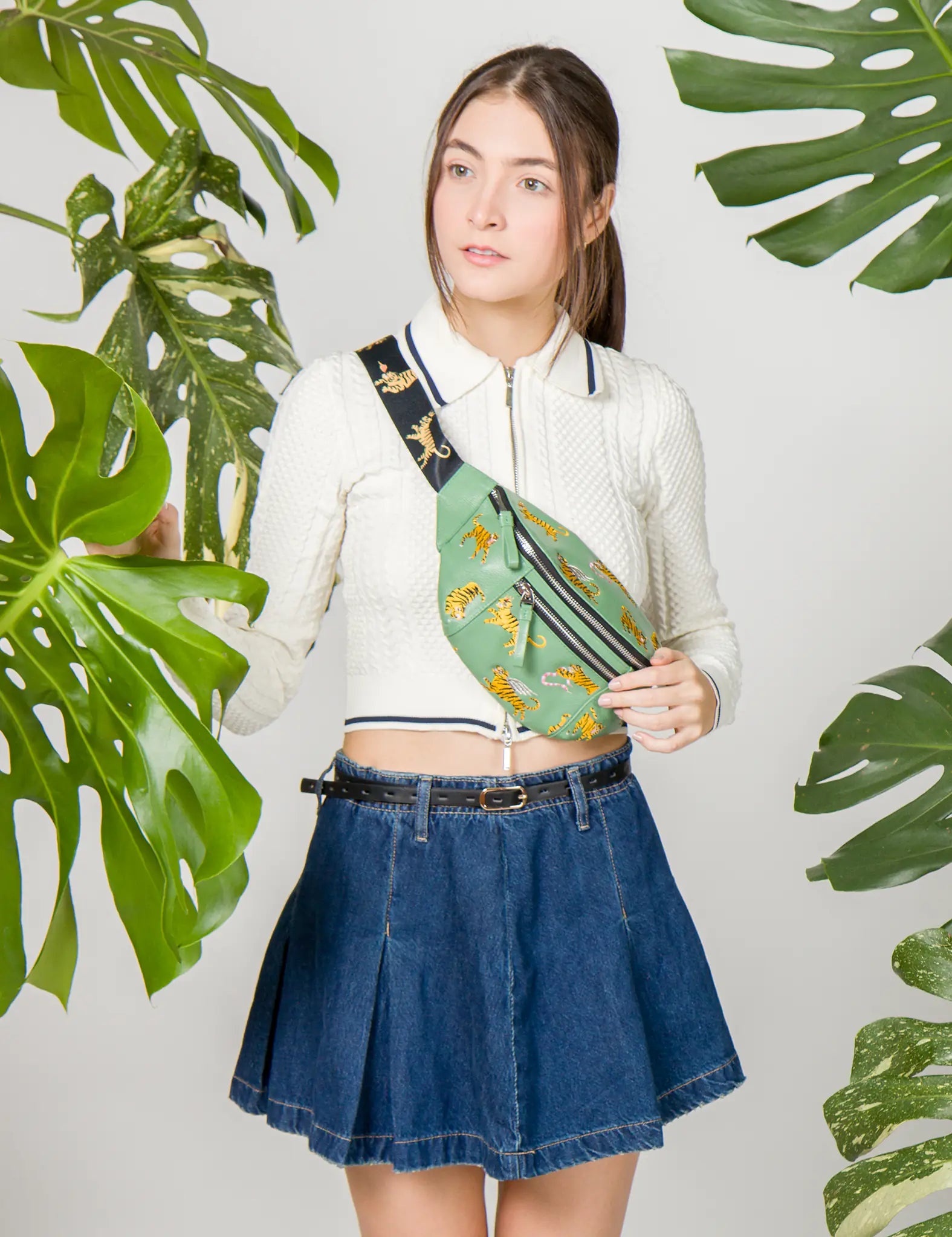 Woman wearing a white knit top and denim skirt poses with a green fanny pack decorated with embroidered tigers, styled crossbody with a wide black strap featuring golden tiger motifs.