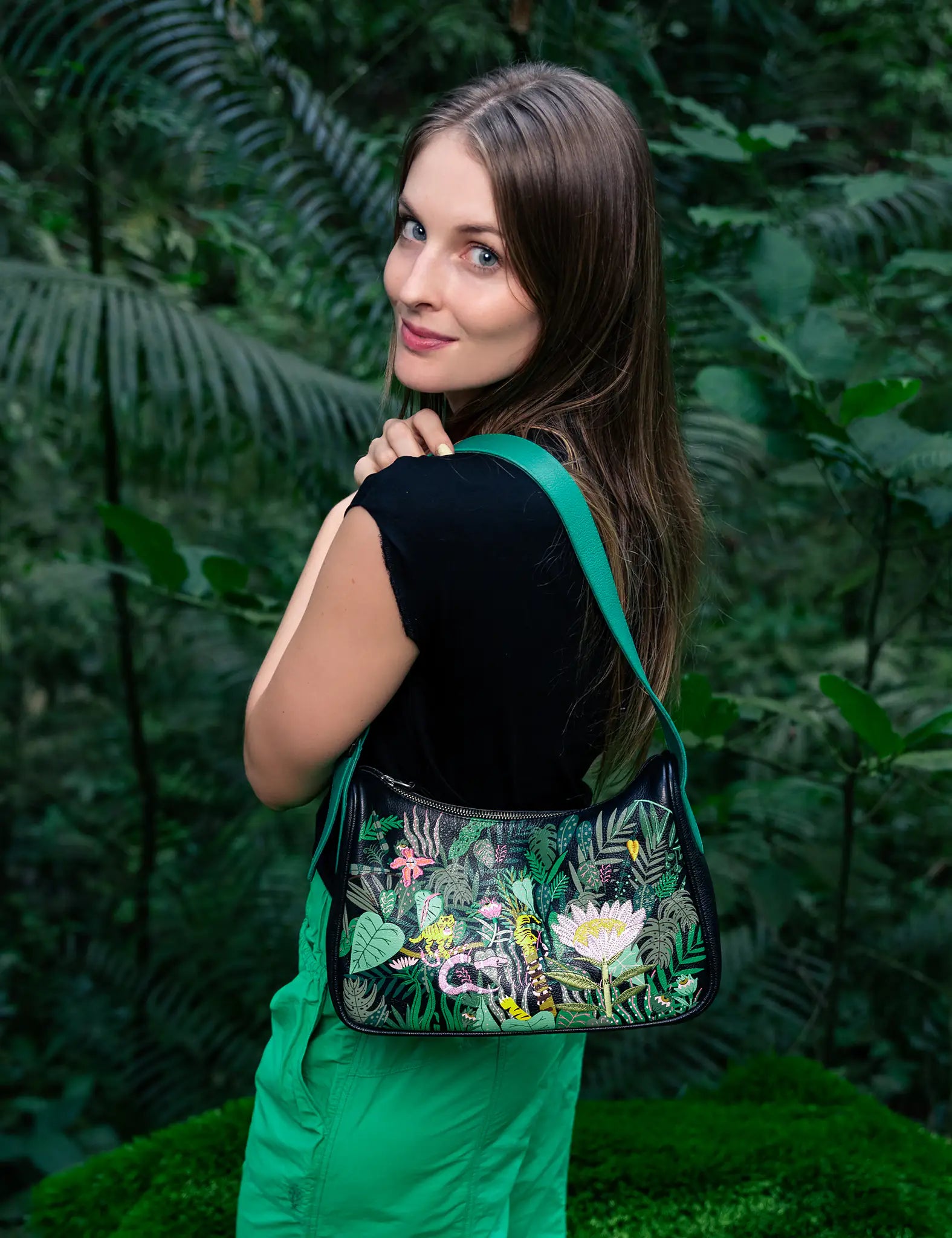 A young woman with long brown hair is standing in a lush, green forest. She is wearing a black sleeveless top and green pants. Over her shoulder, she carries a Vynil Hobo leather handbag by Min & Mon, a NYC-based brand. The handbag features a vibrant, jungle-inspired design with intricate botanical and animal motifs.
