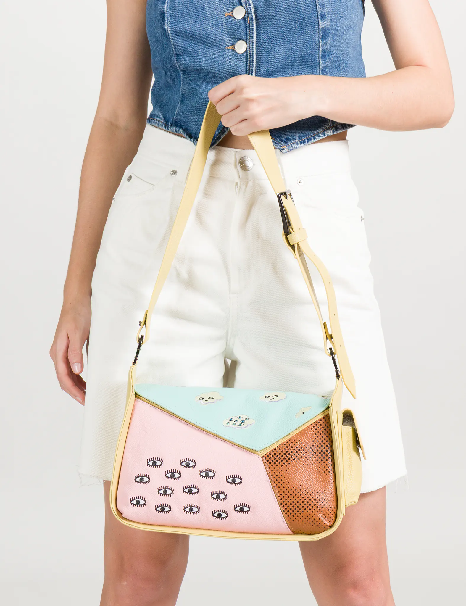 Model in a sleeveless denim top and white shorts holding a color-block shoulder bag with pastel blue, pink, and brown panels, decorated with embroidered eye motifs.